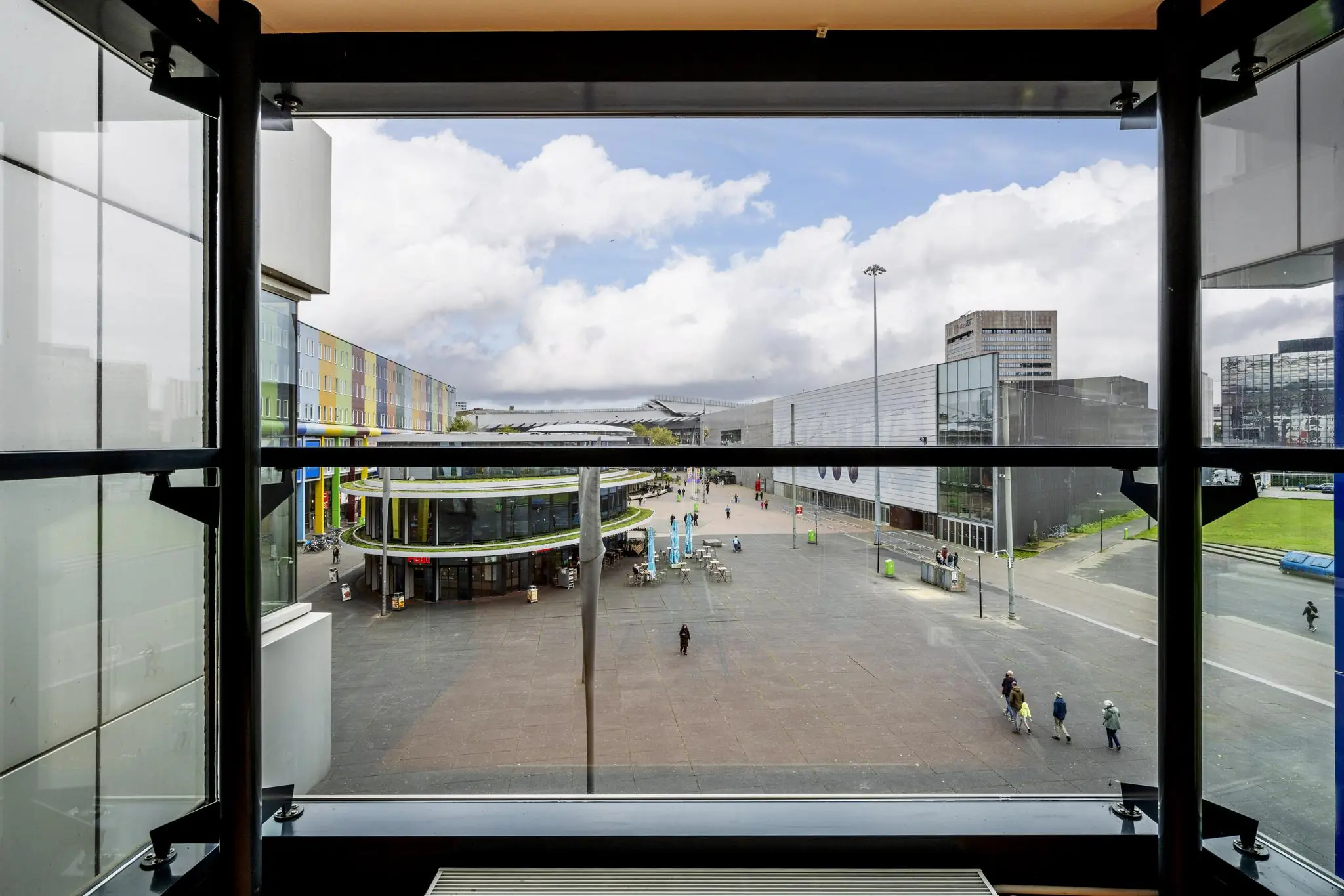 Uitzicht vanaf een raam op de Johan Cruijff Boulevard met moderne gebouwen, wandelend publiek en een plein.