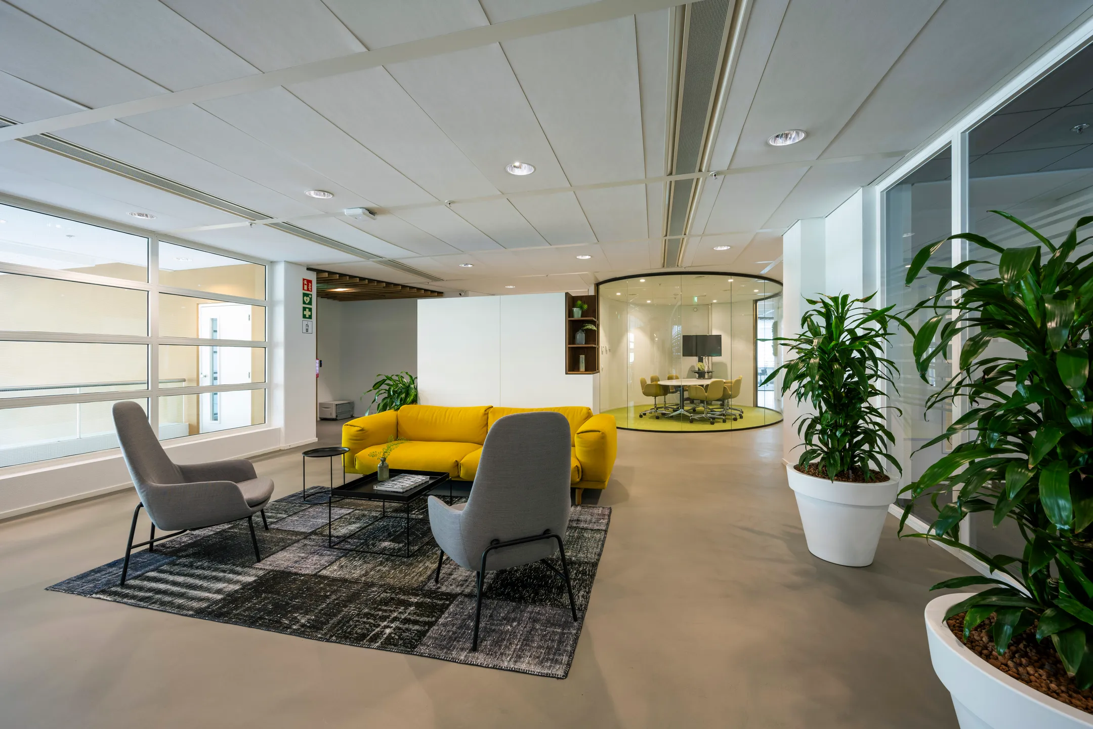 Een moderne kantoorruimte met een gele bank, grijze stoelen, en een glazen vergaderruimte.