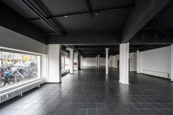 Leegstaand zwart-wit interieur van een ruime commerciële ruimte aan de Muiderstraat met grote ramen en zicht op geparkeerde fietsen buiten.