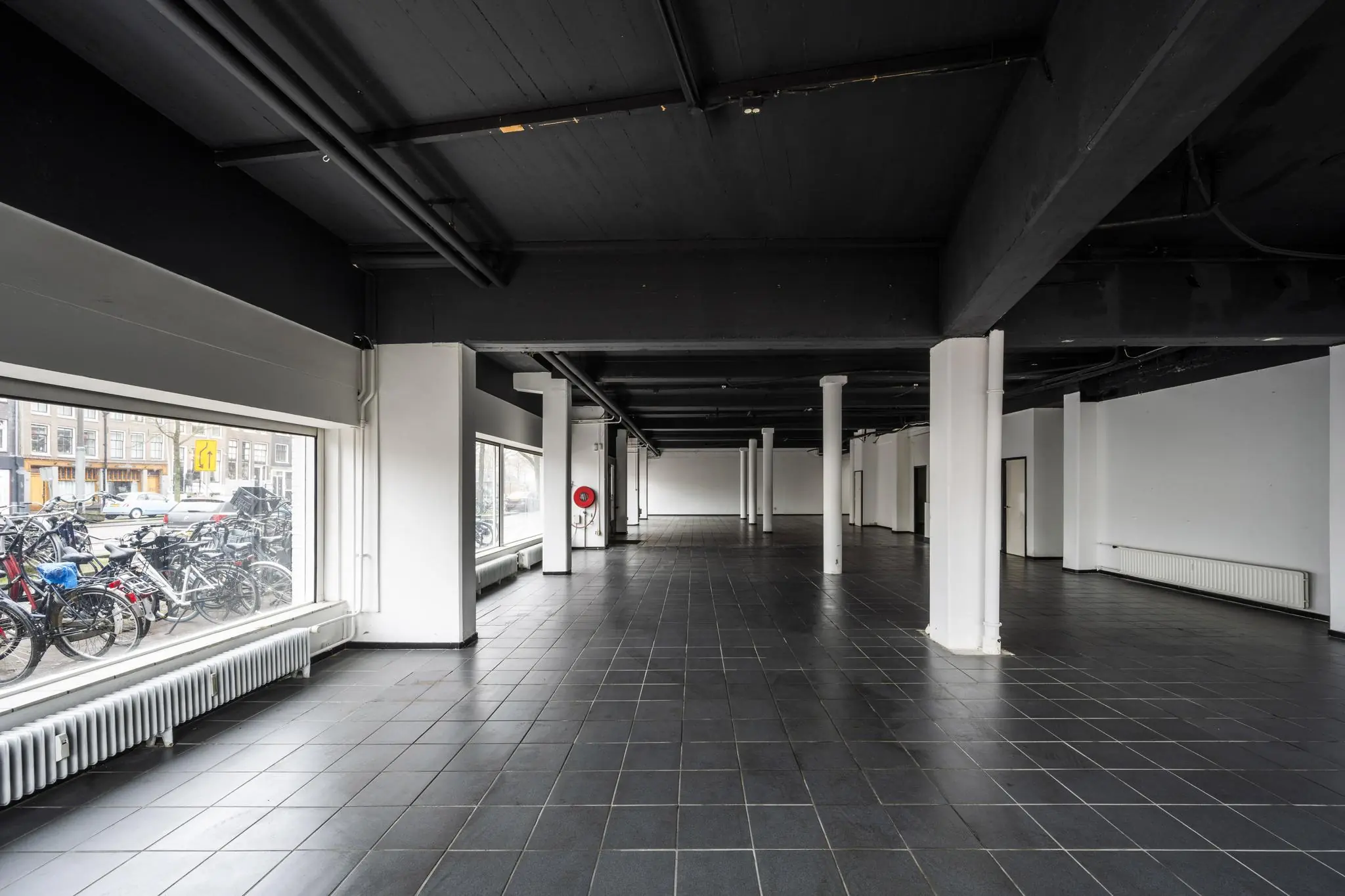 Leegstaand zwart-wit interieur van een ruime commerciële ruimte aan de Muiderstraat met grote ramen en zicht op geparkeerde fietsen buiten.