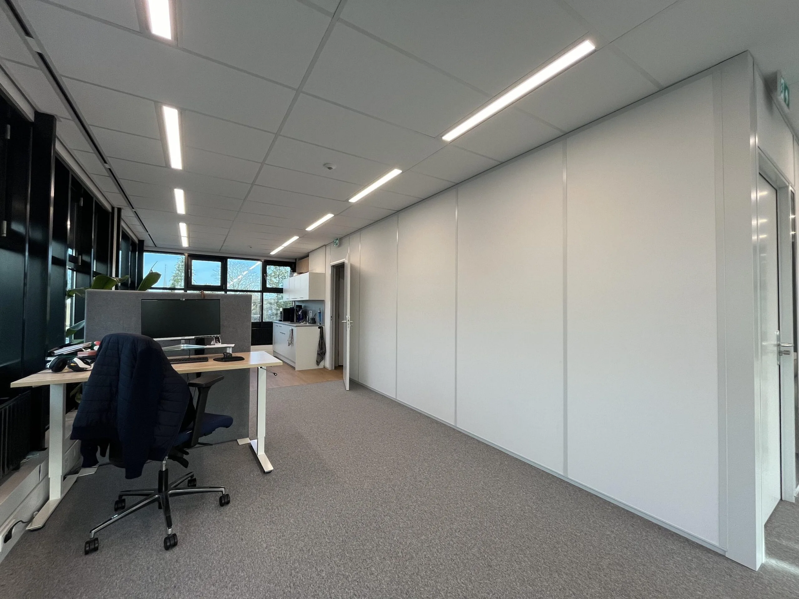 Een moderne kantoorruimte met een bureau, bureaustoel, computer en grote ramen aan de linkerkant.