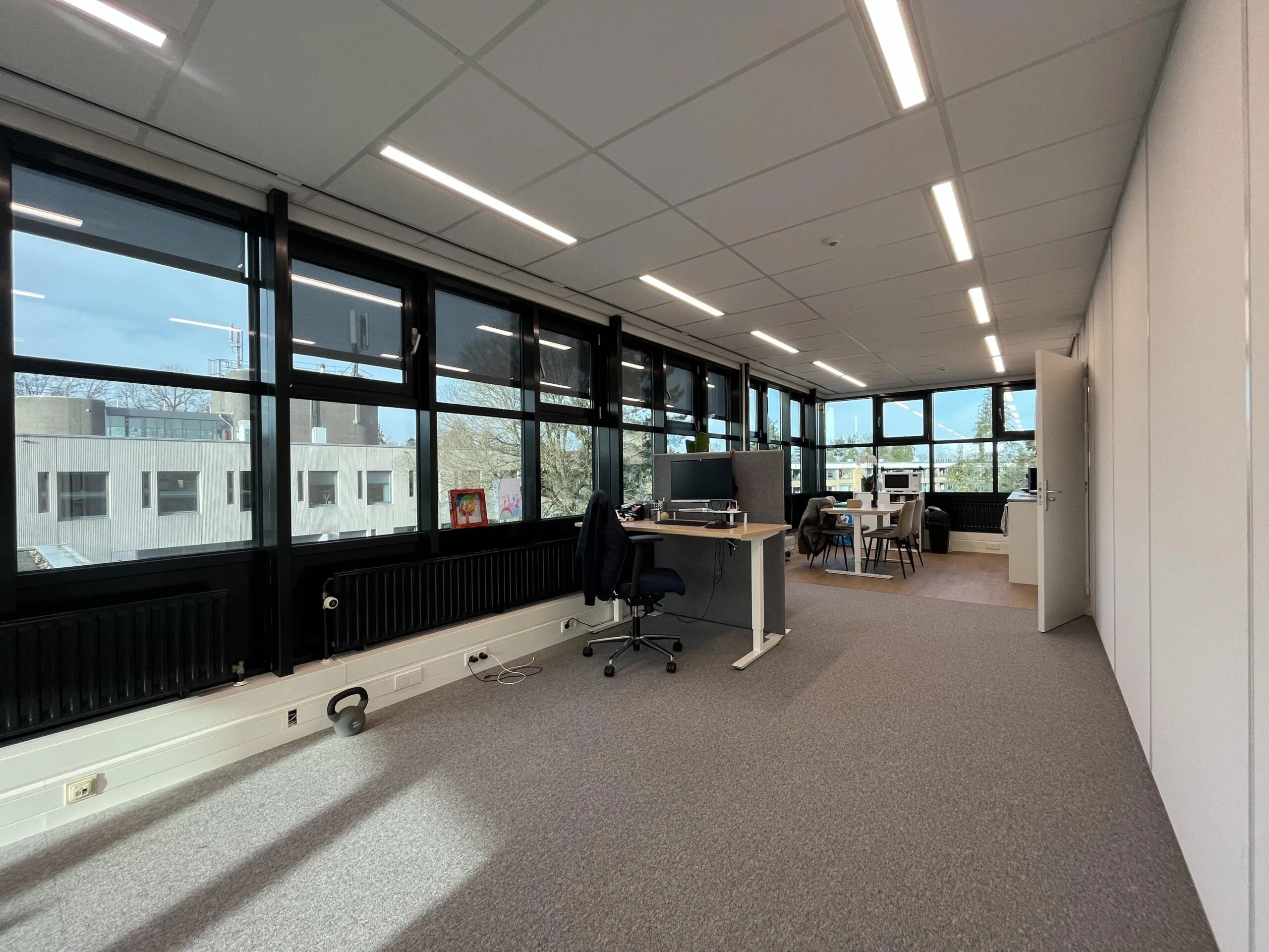 Een ruime, moderne kantoorruimte met bureaus, stoelen en grote ramen die uitzicht bieden op een gebouw.