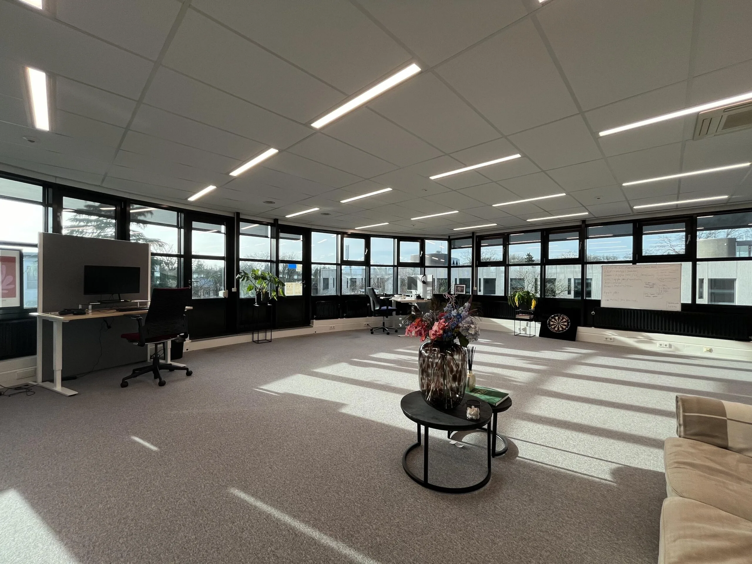 Een ruime, moderne kantoorruimte met veel natuurlijk licht, een bureau met stoel, planten en een dartbord.