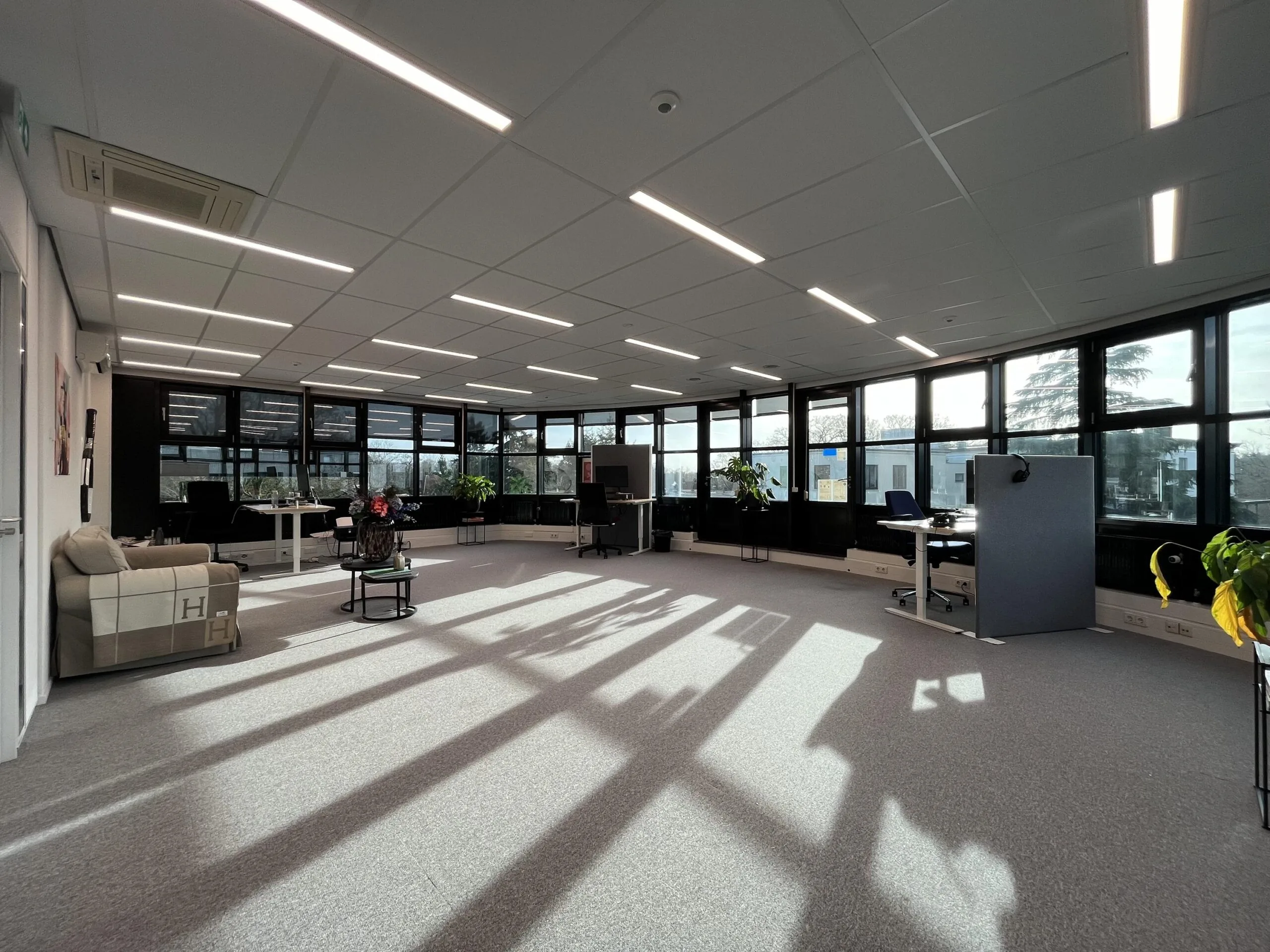 Een ruime, moderne kantoorruimte met veel natuurlijk licht, voorzien van bureau's, planten en een zitgedeelte.