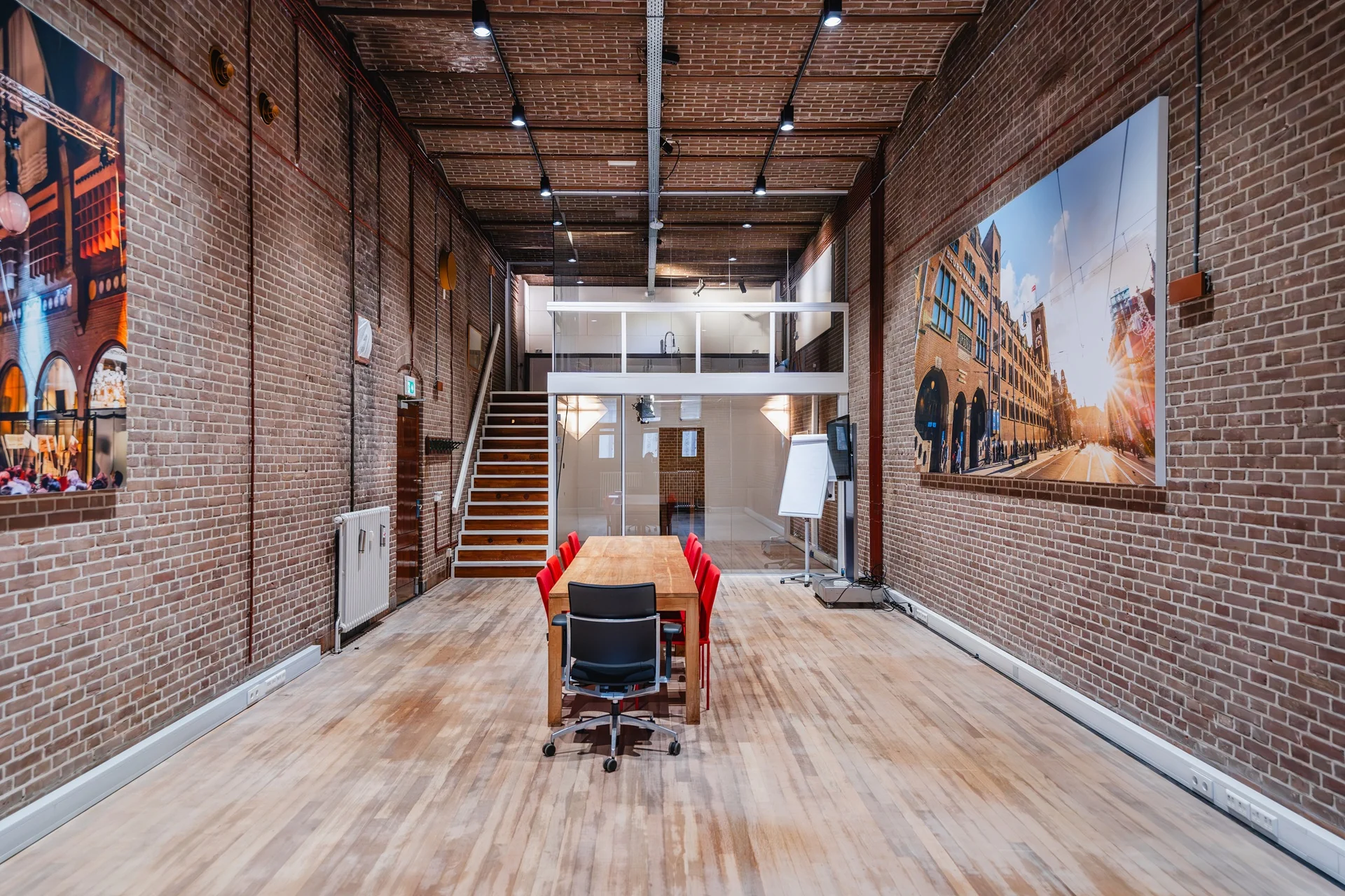 Vergaderruimte met bakstenen muren, houten vloer, een lange tafel met rode stoelen en een grote foto van de Damrak in Amsterdam aan de muur.
