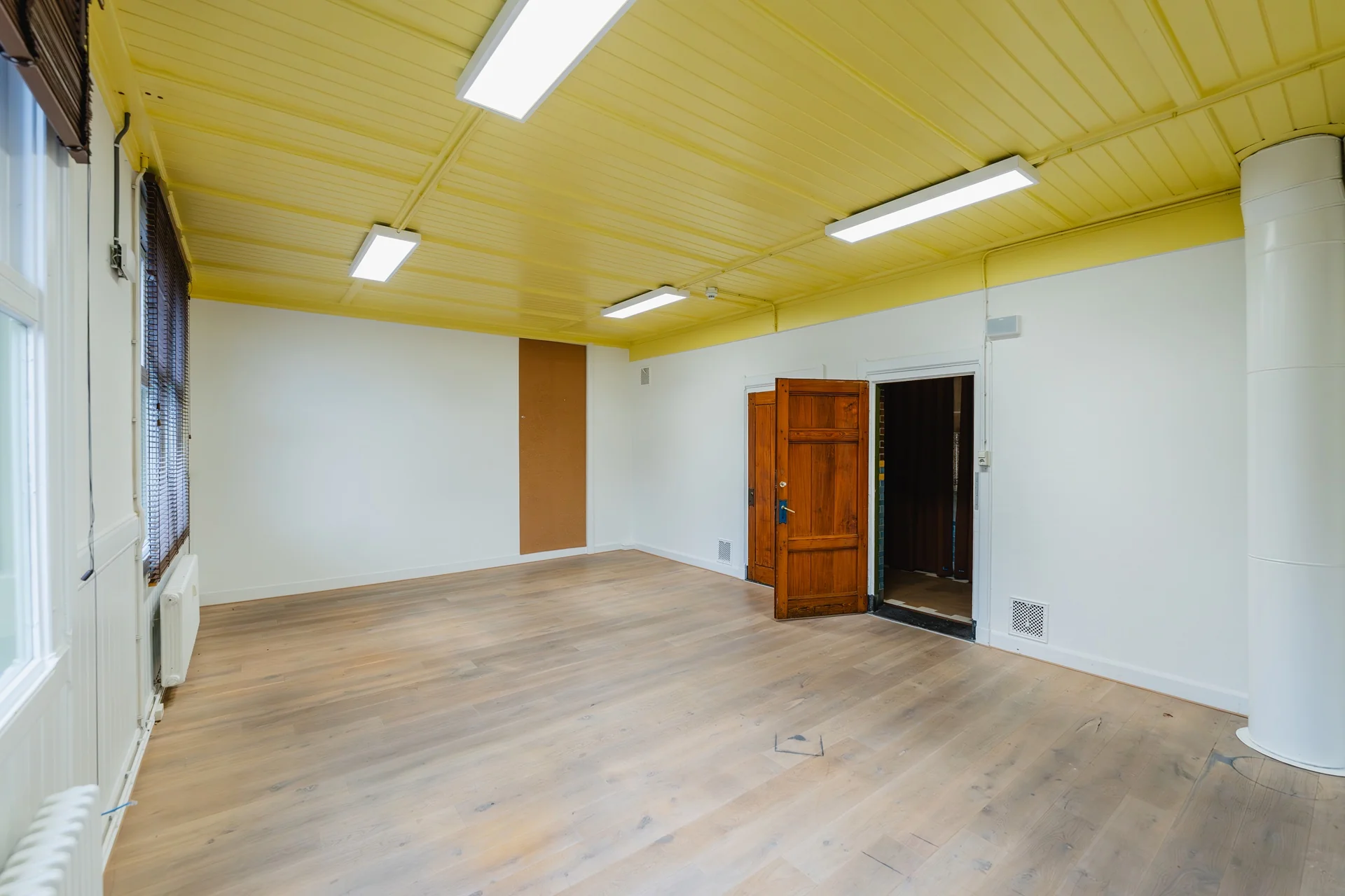 Lege kamer aan het Damrak met gele plafondpanelen, houten vloer en open houten deur.