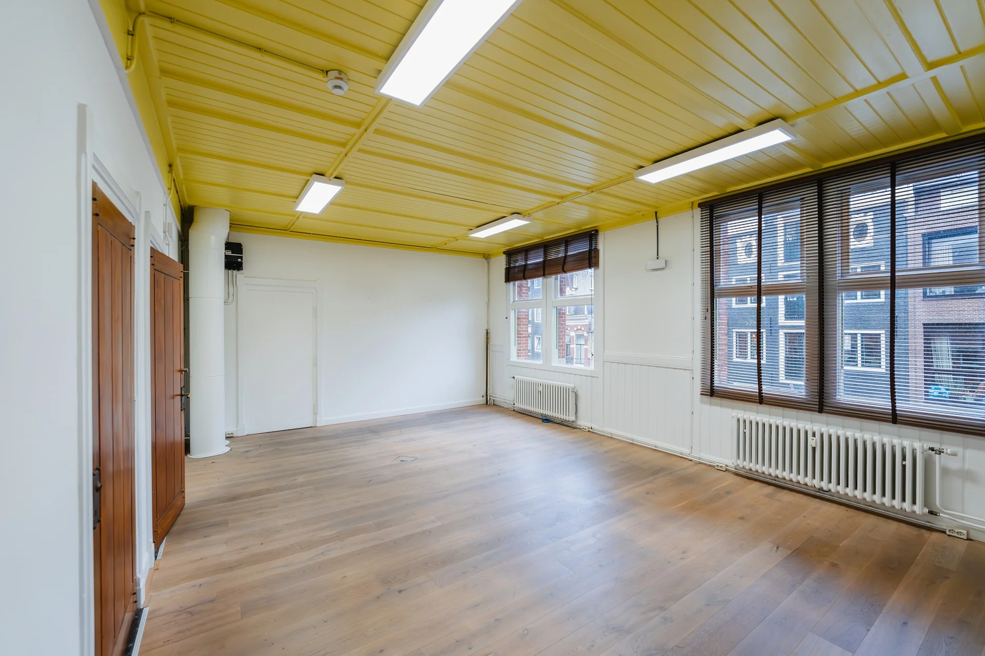 Lichte, lege ruimte aan het Damrak met houten vloer, geel plafond en grote ramen met jaloezieën.