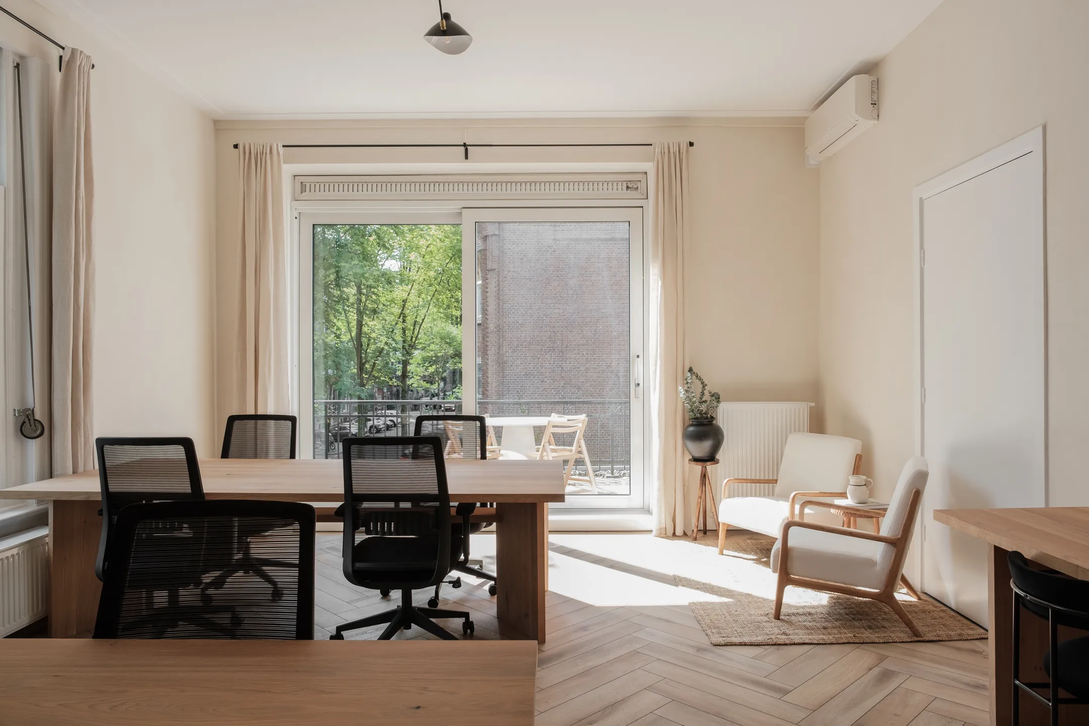 Een lichte werkkamer met houten bureau, meerdere stoelen en een deur naar een balkon met uitzicht op bomen.