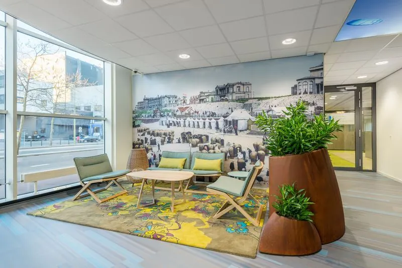 Wachtgedeelte met moderne stoelen, kleurrijk vloerkleed en een grote muurprint van het Spui in Den Haag.