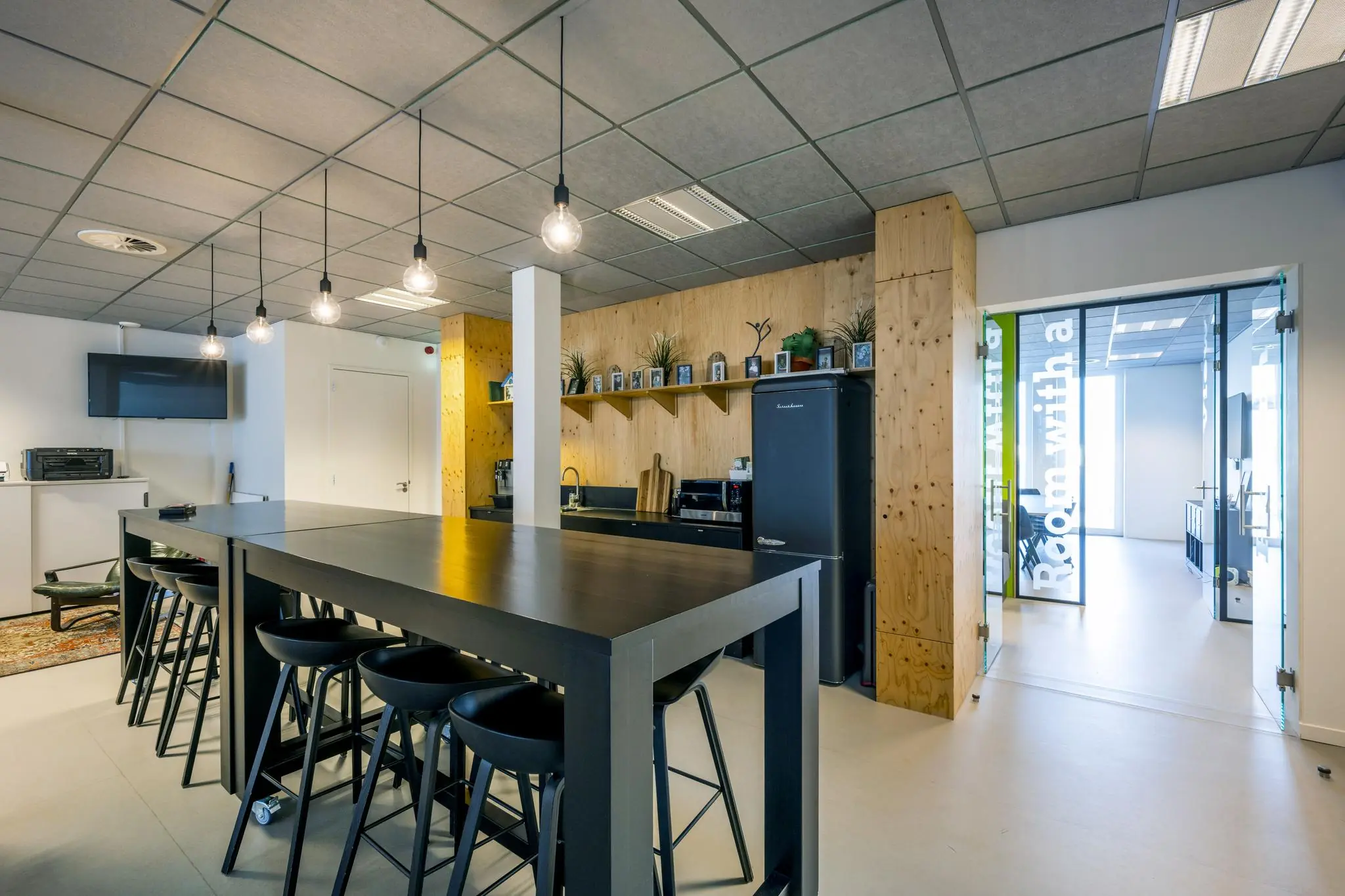 Moderne kantoorkeuken aan de De Ruijterkade met een lange hoge eettafel, hanglampen en een houten wand met open schappen.