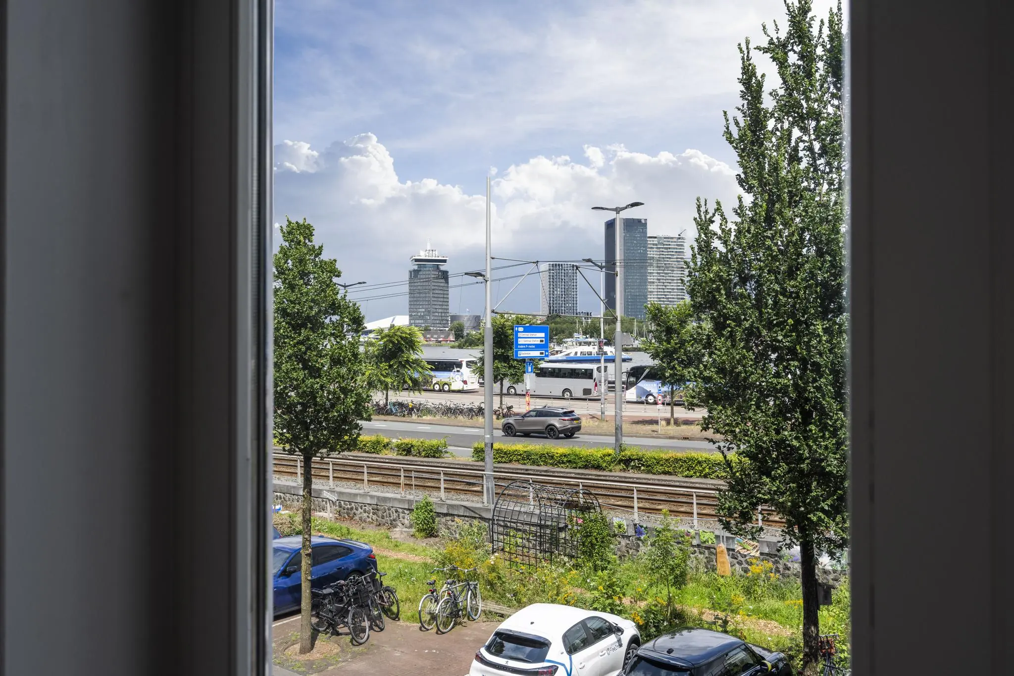 Uitzicht vanuit een raam op De Ruijterkade in Amsterdam met trambaan, auto's, fietsen en moderne hoogbouw op de achtergrond.