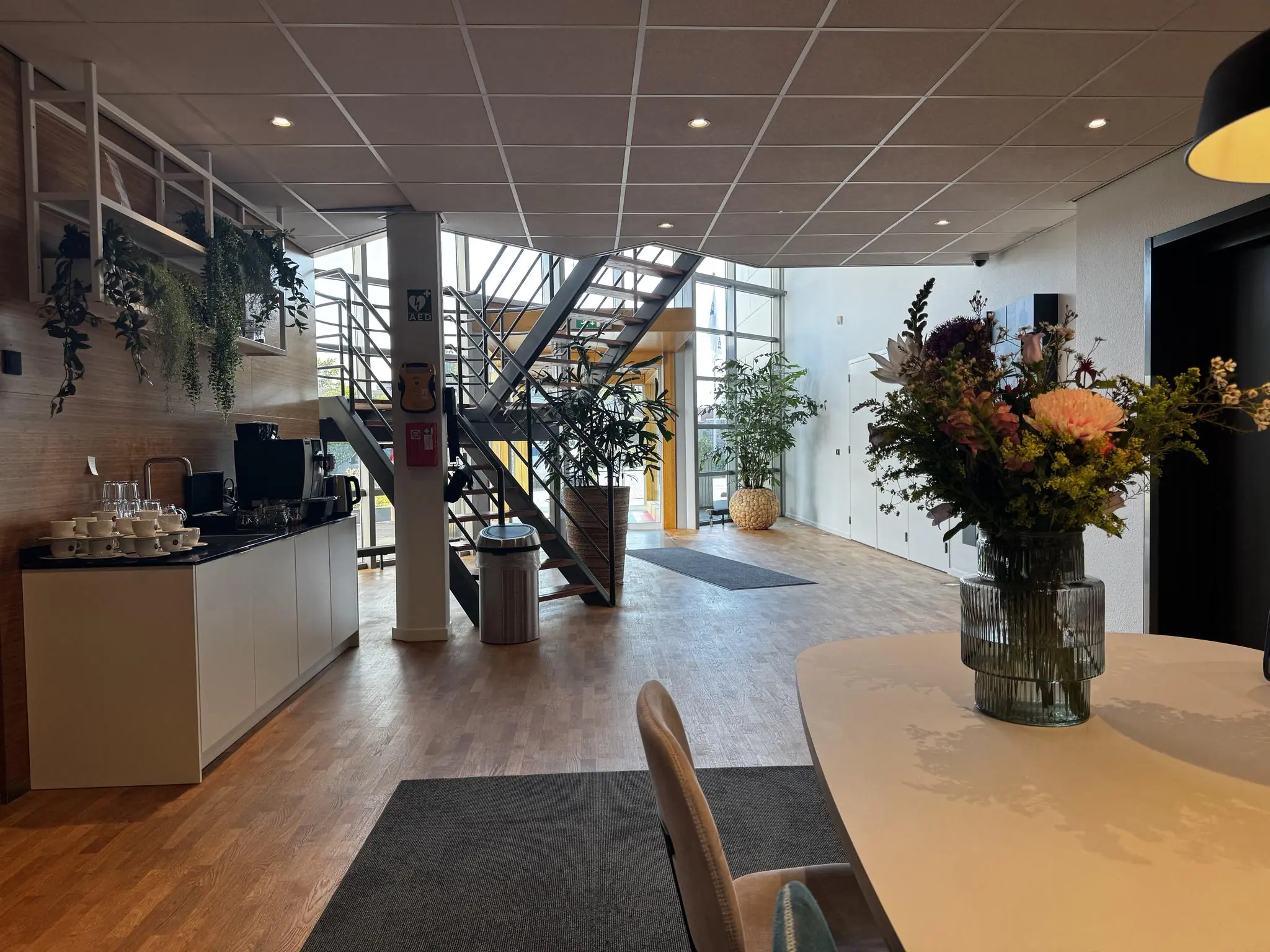 Moderne kantoorentree met een koffiecorner, bloemen op tafel en een trap naar de bovenverdieping bij Softwareweg.