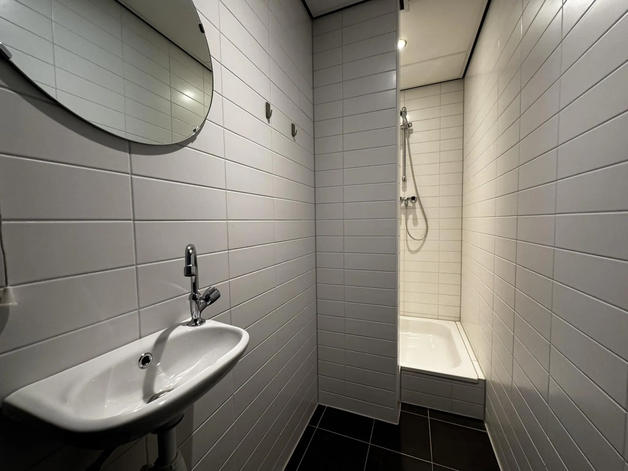 Badkamer met witte wandtegels, een kleine wastafel, ronde spiegel en een douchegedeelte met douchekop.