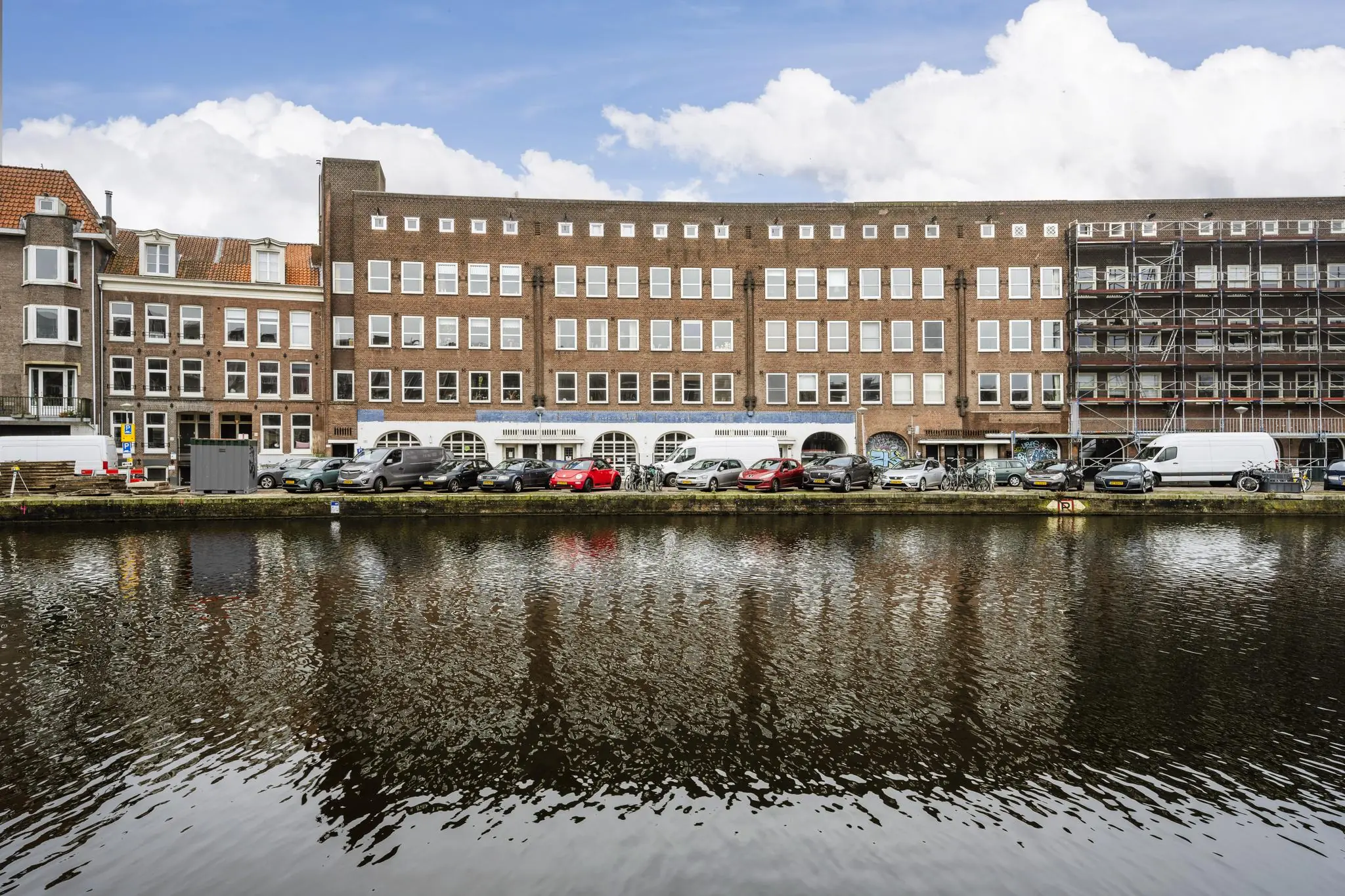Rij met geparkeerde auto's langs de Baarsjesweg in Amsterdam, met karakteristieke bakstenen gebouwen en uitzicht op het kanaal.