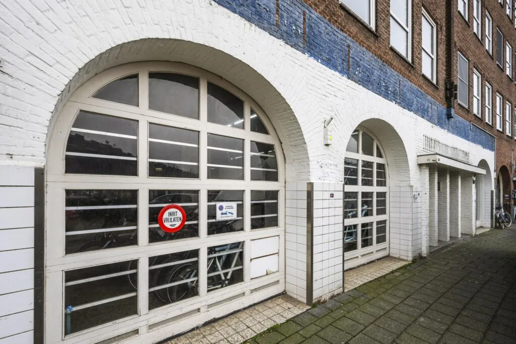 Witgepleisterde boogvormige garagedeuren aan de Baarsjesweg met fietsen zichtbaar achter glas en een bordje “Inrit vrijlaten”.