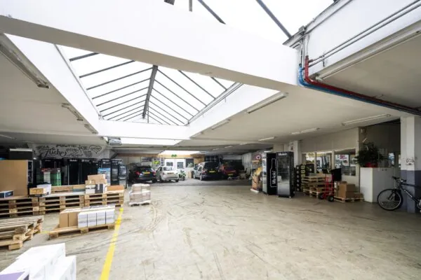 Magazijnruimte aan de Baarsjesweg met geparkeerde auto's, stapels dozen op pallets en een glazen dak voor natuurlijk licht.