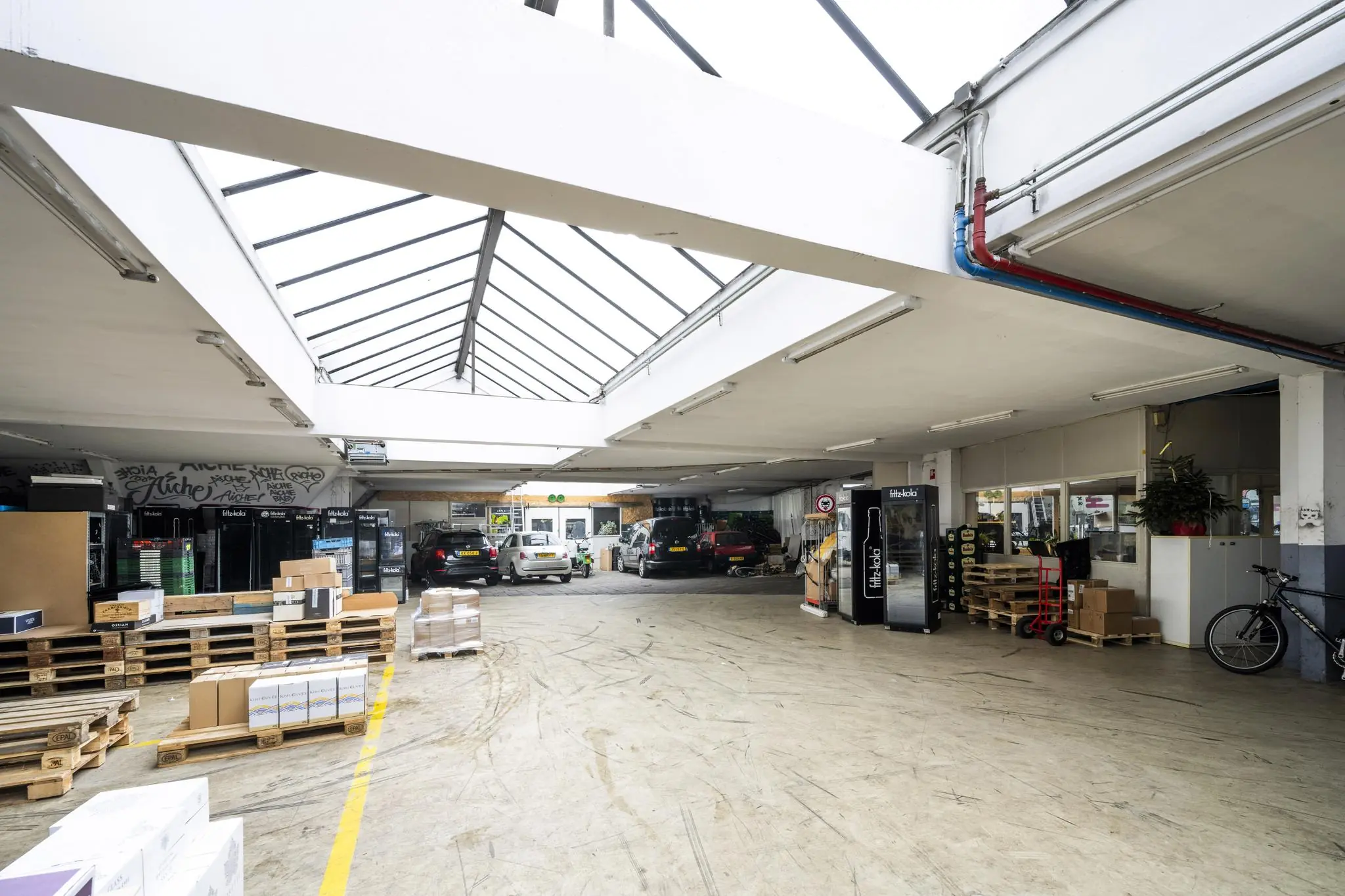 Magazijnruimte aan de Baarsjesweg met geparkeerde auto's, stapels dozen op pallets en een glazen dak voor natuurlijk licht.