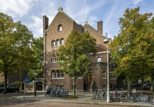 Monumentaal bakstenen pand aan het Emmaplein in Amsterdam, omringd door bomen en geparkeerde fietsen.