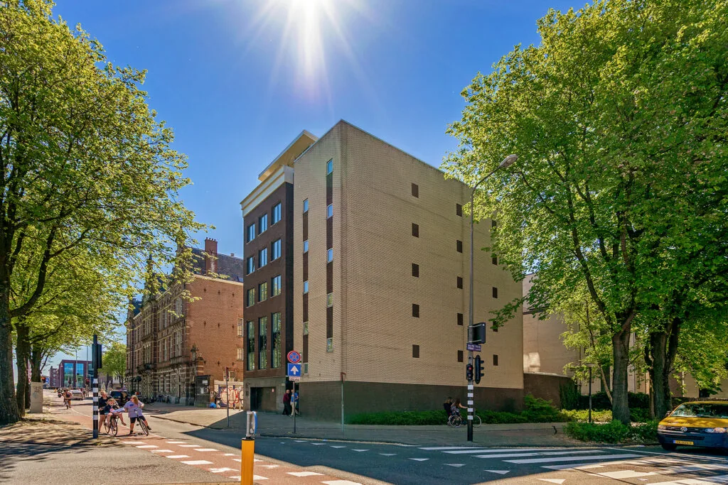 Zonnige straatzicht op de Prins Hendrikkade in Amsterdam met moderne en historische gebouwen, fietsers en groen langs de weg.