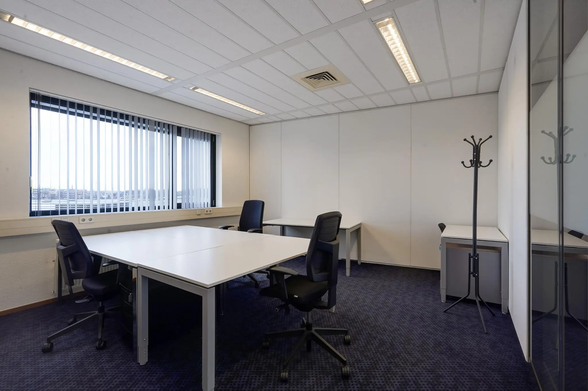 Kantoorruimte aan de Laan van ’s-Gravenmade met bureaus, bureaustoelen, een kapstok en een groot raam met verticale lamellen.