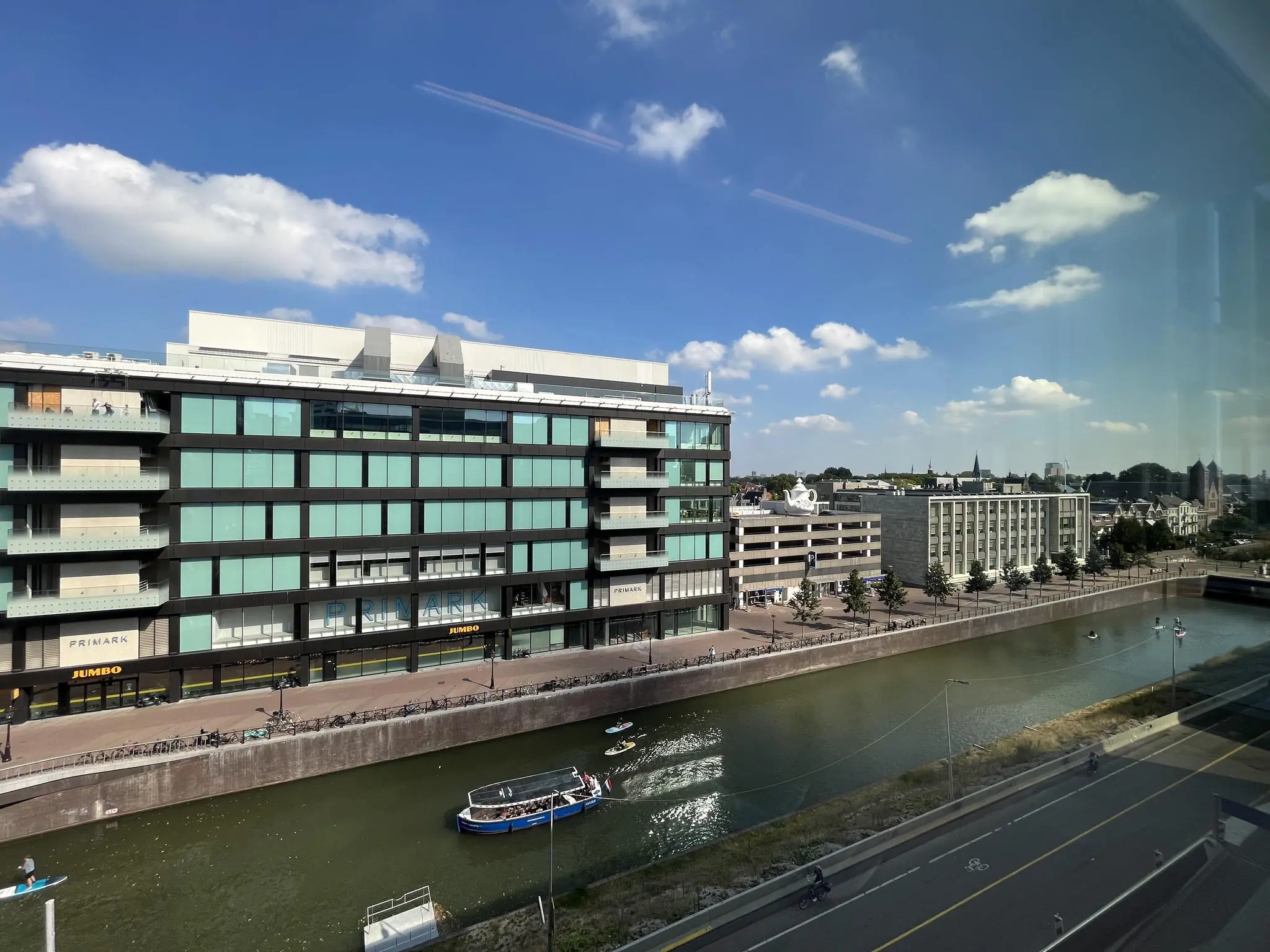 Uitzicht op de Catharijnesingel in Utrecht met een modern gebouw, winkels zoals Primark en Jumbo, en boten op het water.