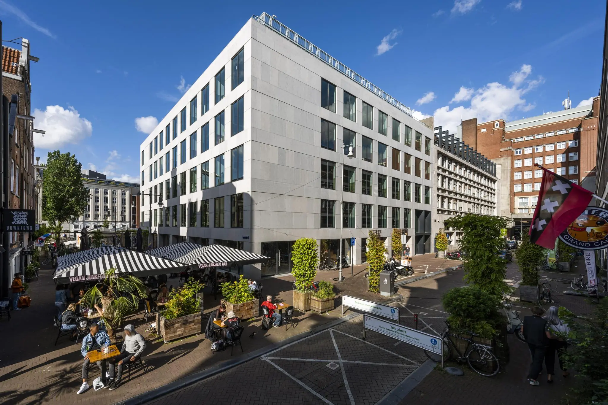 Terras van Vegan Junk Food Bar aan de Singel in Amsterdam met moderne kantoorgebouwen en voetgangers in de zon.