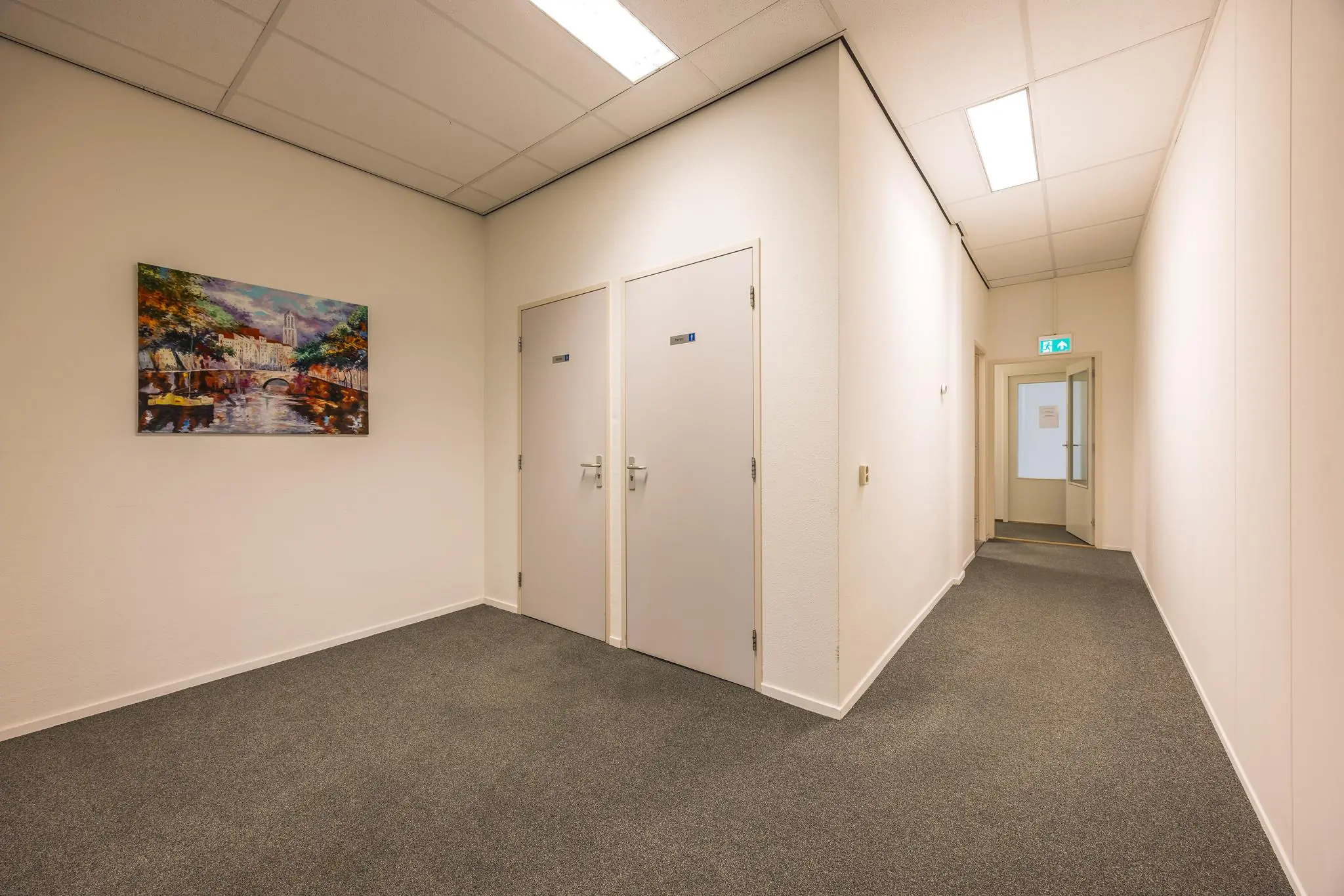 Verlichte gang met twee gesloten deuren, een schilderij aan de muur en een open deur naar een andere ruimte aan het einde van de gang.