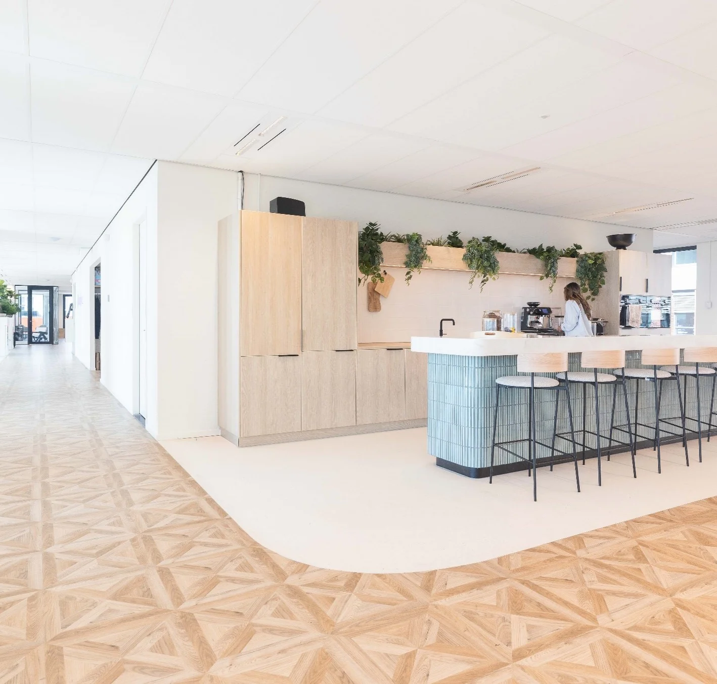 Moderne kantoorkeuken aan de Mt. Lincolnweg met een houten vloer, een bar met krukken en een vrouw bij het koffieapparaat.