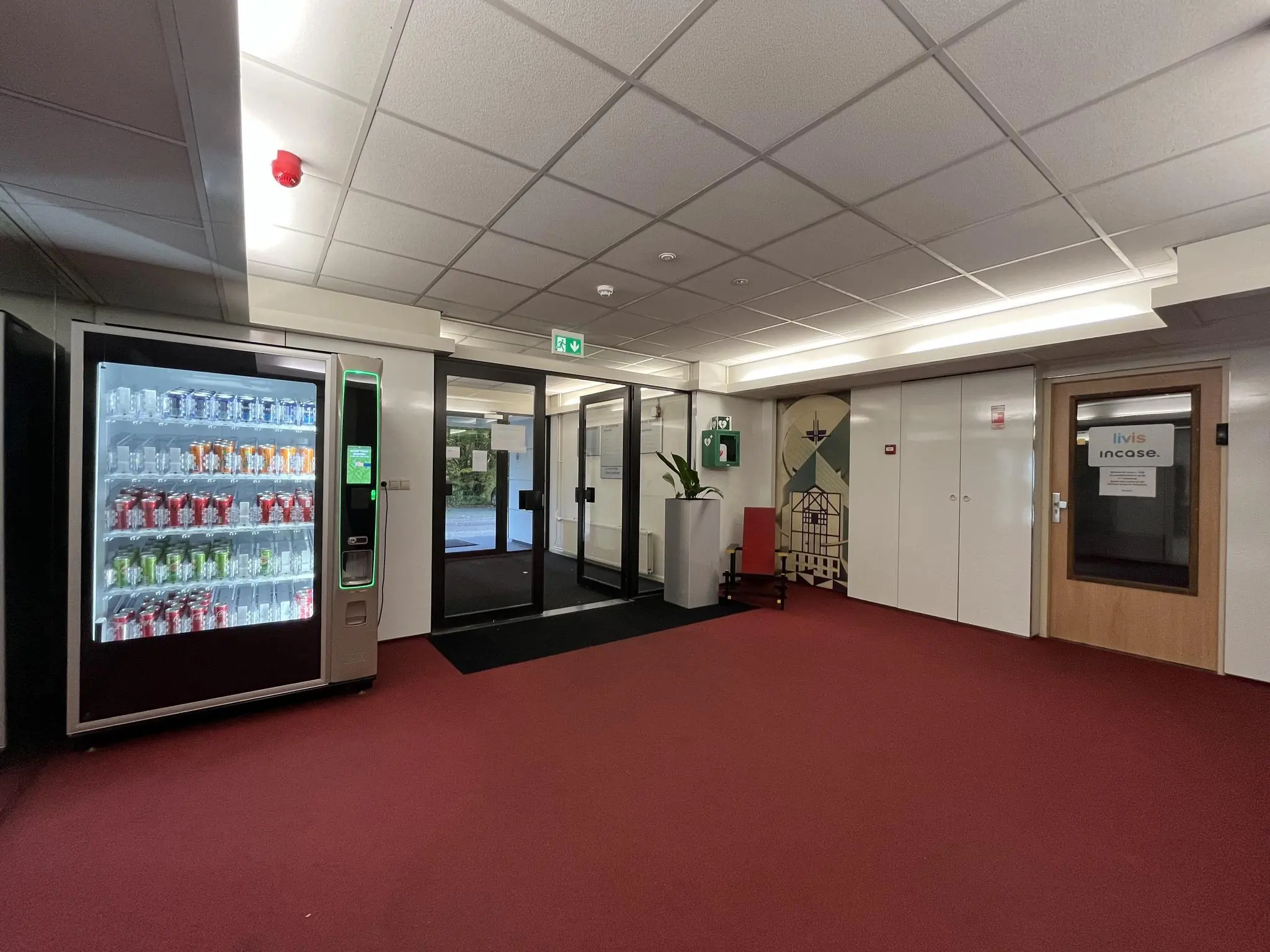 Hal met rode vloerbedekking, frisdrankautomaat, glazen deuren en een deur met het bordje "Livis incase".