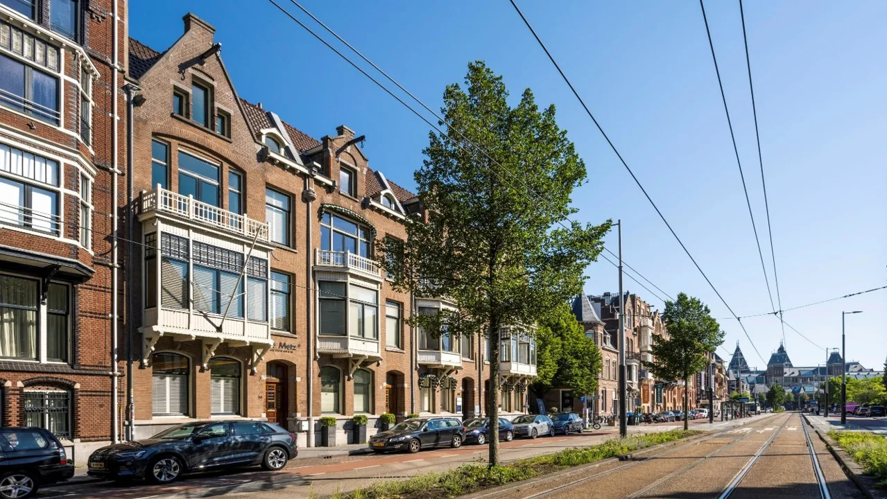 Straatbeeld van de Paulus Potterstraat in Amsterdam met karakteristieke herenhuizen, geparkeerde auto's en tramrails onder een blauwe lucht.