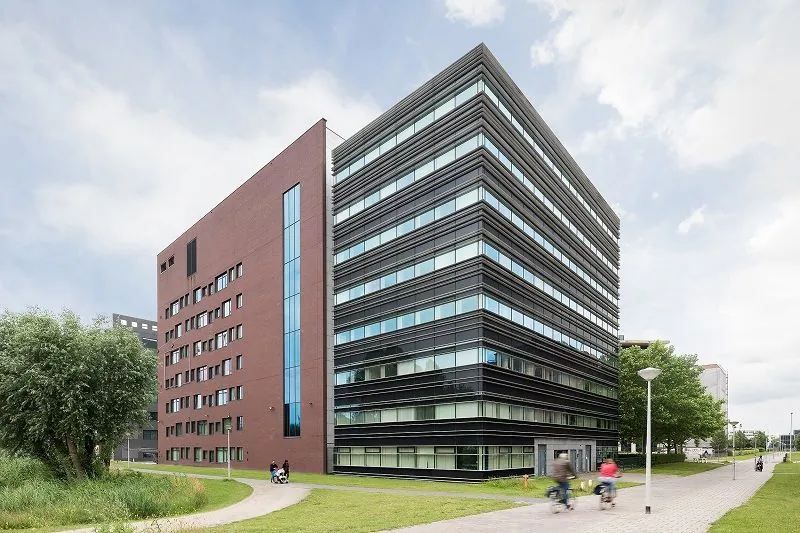 Een modern kantoorgebouw met een rode en zwarte gevel en een pad met fietsers op de voorgrond.