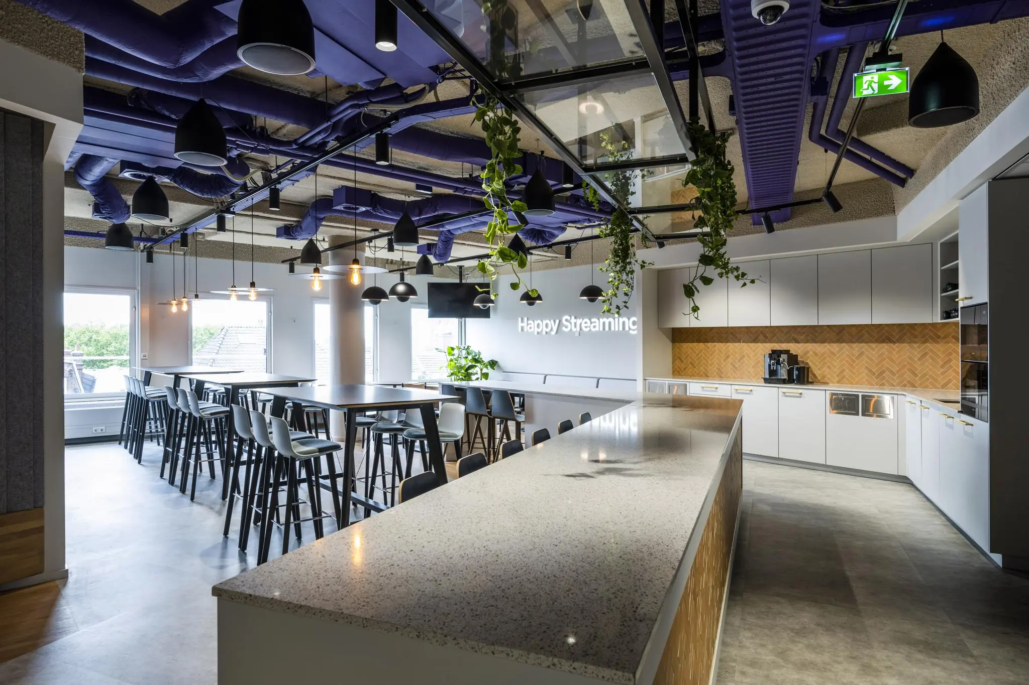 Moderne kantoorkeuken aan de Vijzelgracht met een lange bar, hoge zitplaatsen en het verlichte opschrift "Happy Streaming".