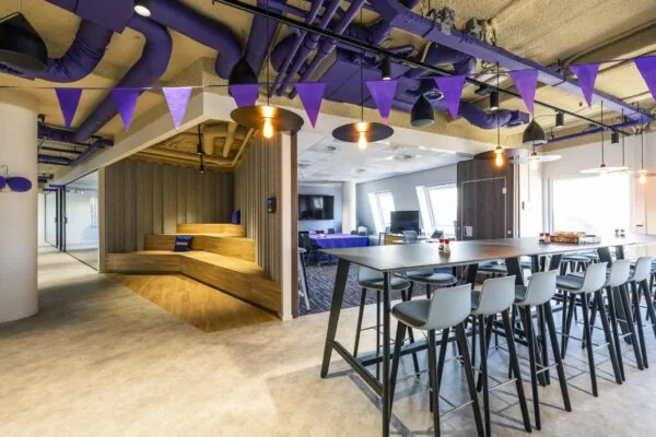 Modern ingerichte kantoorruimte aan de Vijzelgracht met hoge tafels, loungehoek van hout, paarse accenten en speelse plafonddecoratie.