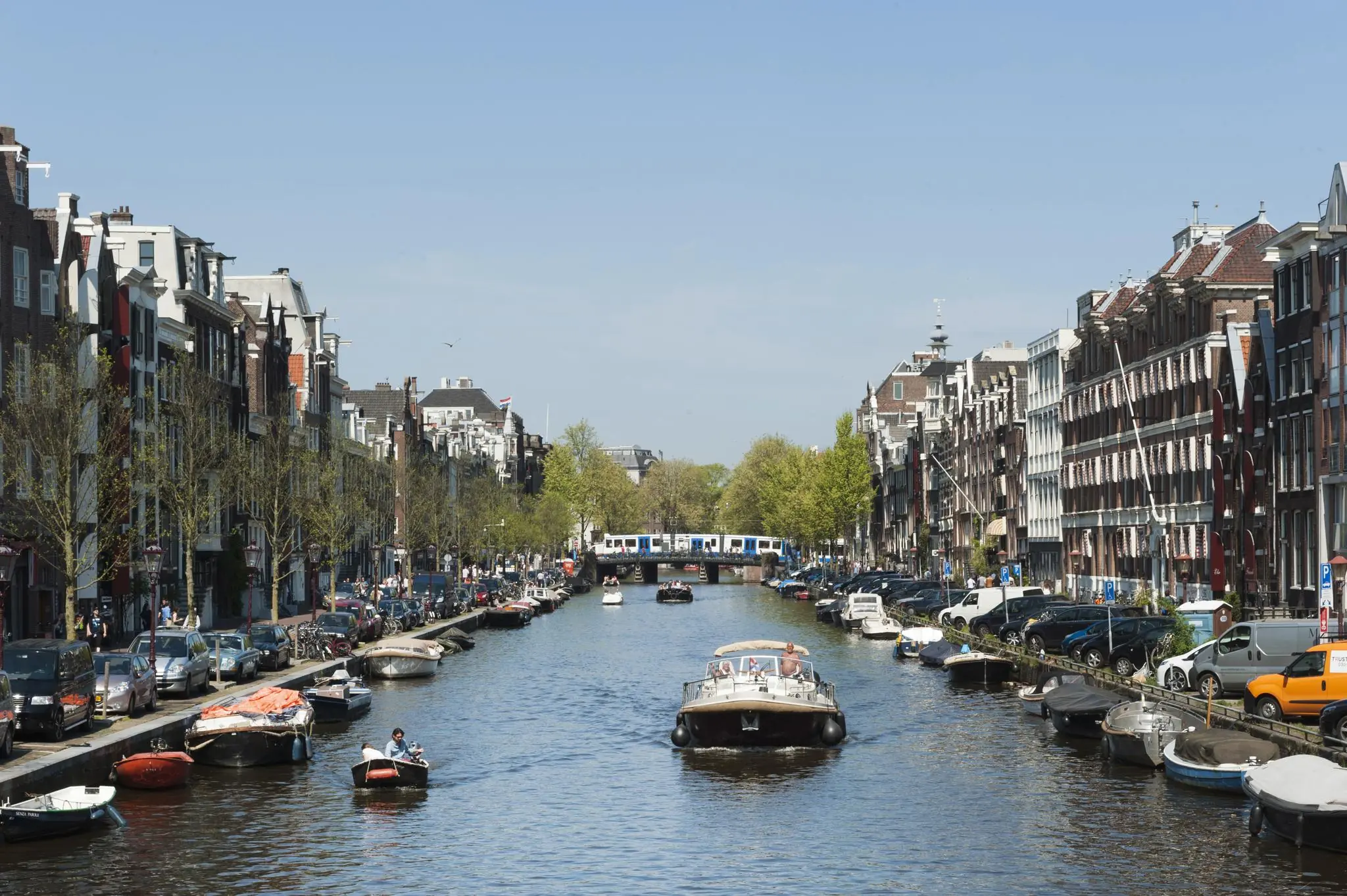 Zicht op de Vijzelgracht in Amsterdam met boten op het water, geparkeerde auto's langs de kant en een metro die over de brug rijdt.