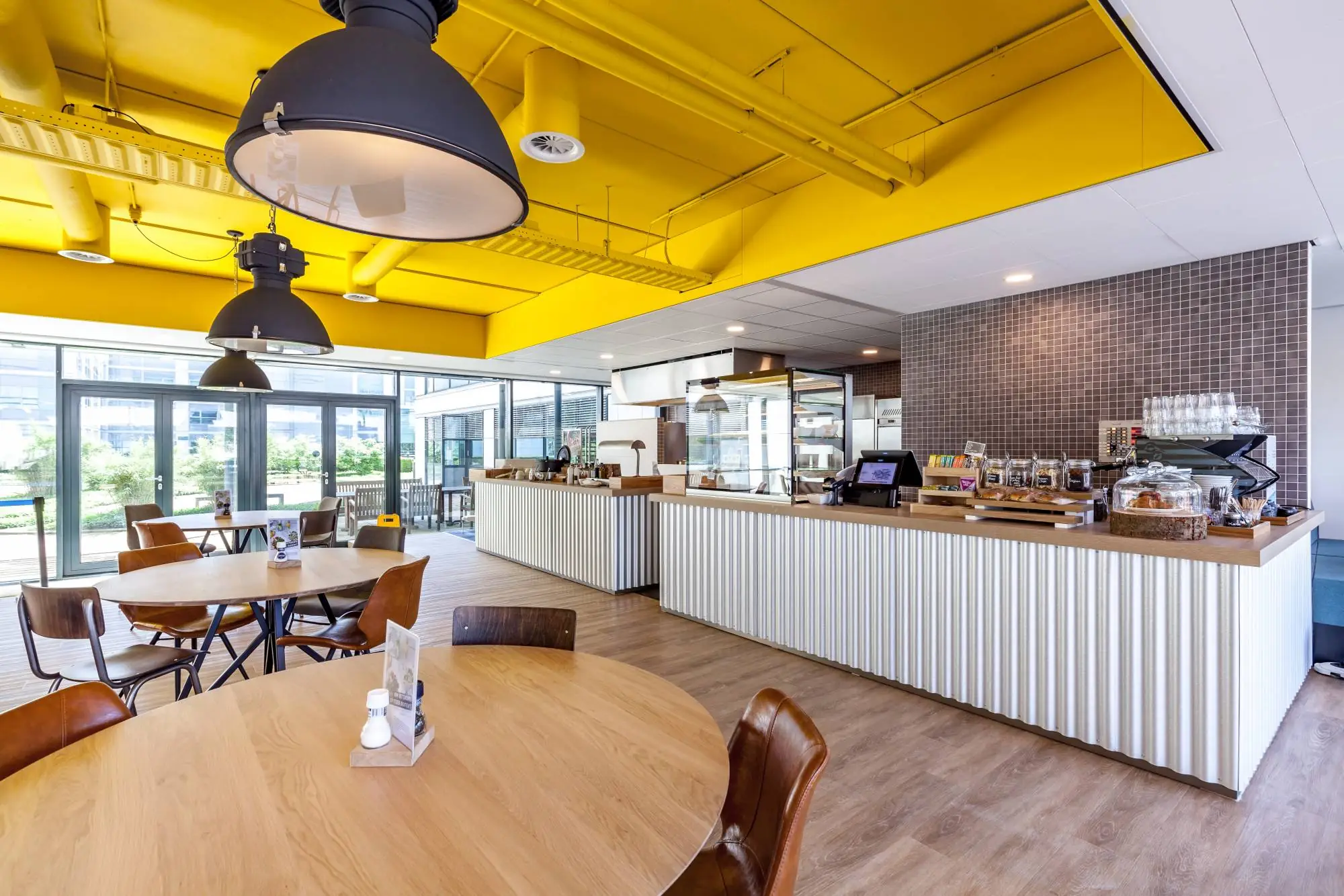 Een modern café-interieur met houten tafels en een gele plafondbalk, naast een balie met etenswaren.