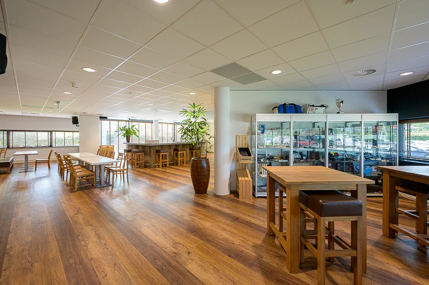 Modern lounge area at Laan van Barcelona with wooden flooring, tables, trophy display cabinets, and large windows providing natural light.