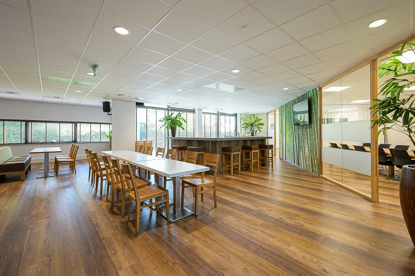 Modern communal dining area at Laan van Barcelona with wooden flooring, tables, chairs, and large windows letting in natural light.