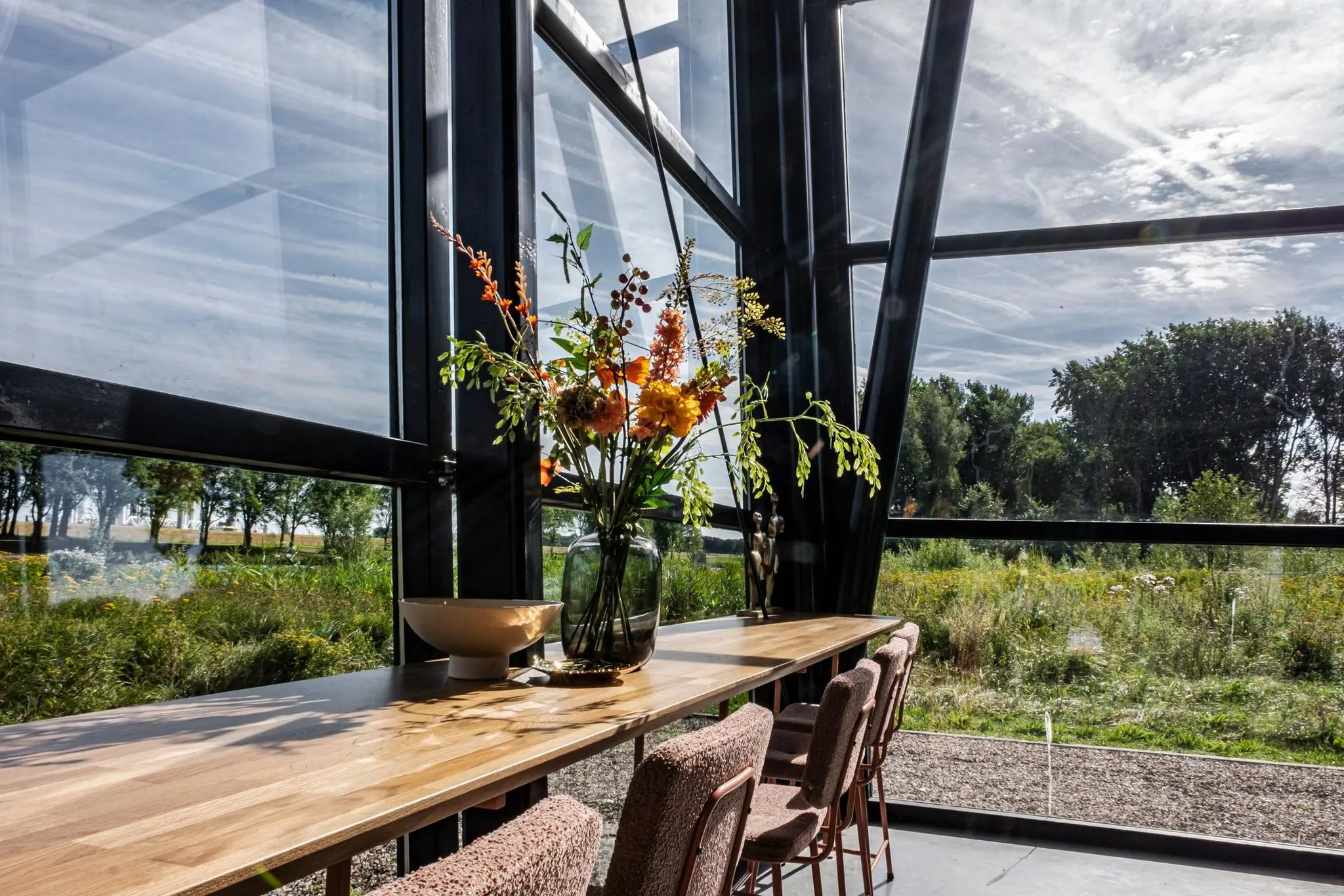 Lange houten eettafel met bloemenvaas in een modern glazen paviljoen met uitzicht op groen landschap.