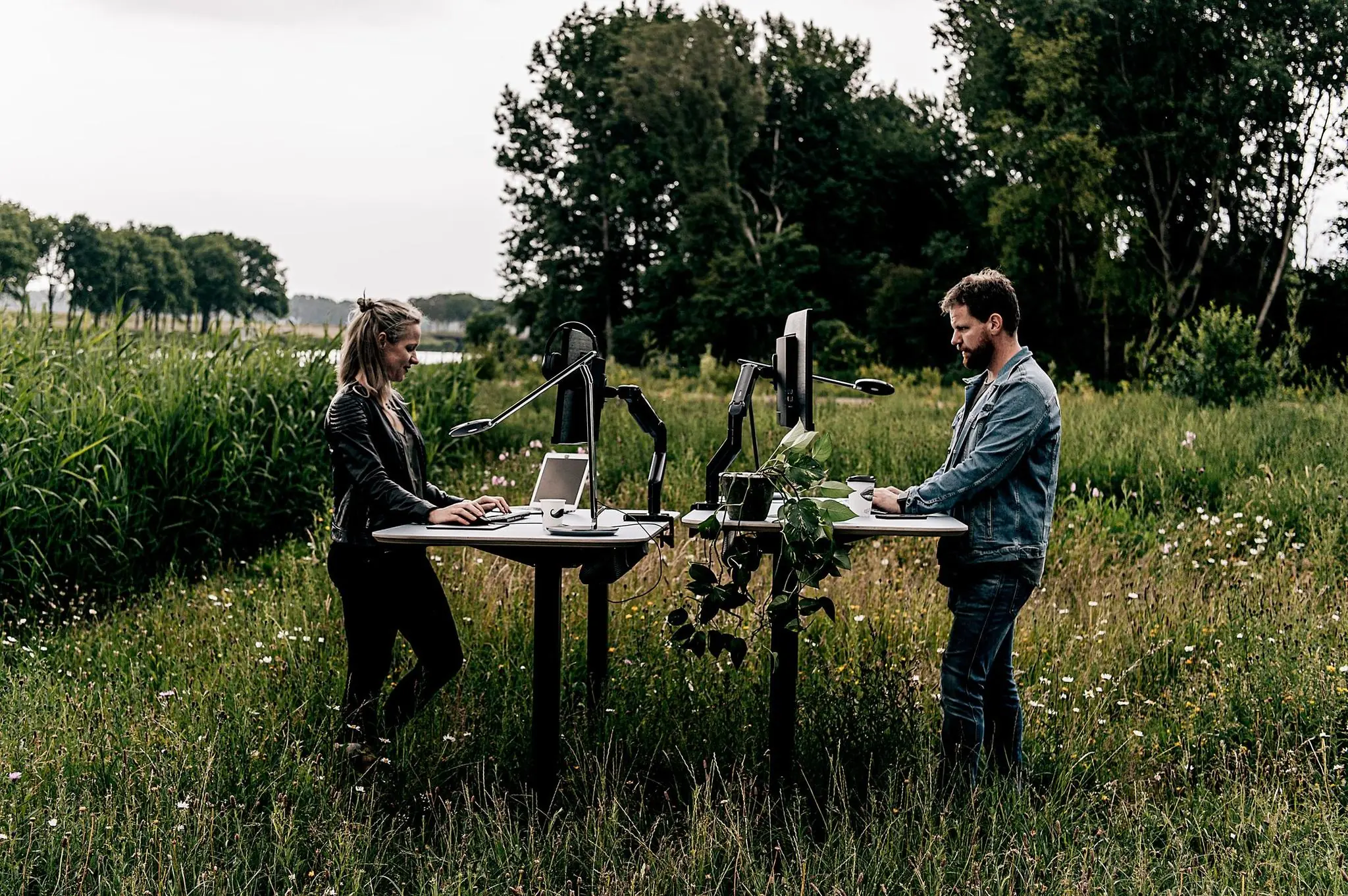 Twee mensen werken op computers aan sta-bureaus in een groen weiland met bomen op de achtergrond.