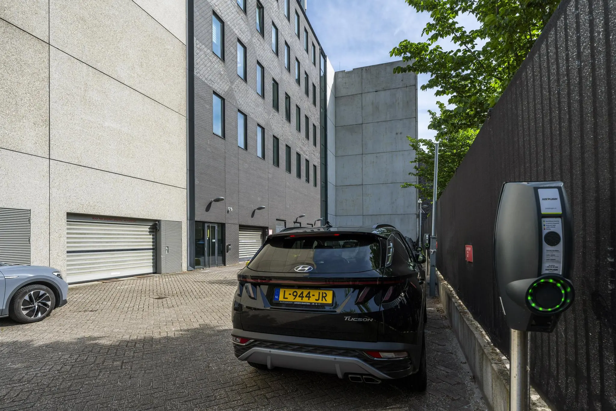 Een zwarte Hyundai Tucson staat geparkeerd en wordt opgeladen bij een laadpaal aan de Krijn Taconiskade naast een modern kantoorgebouw.