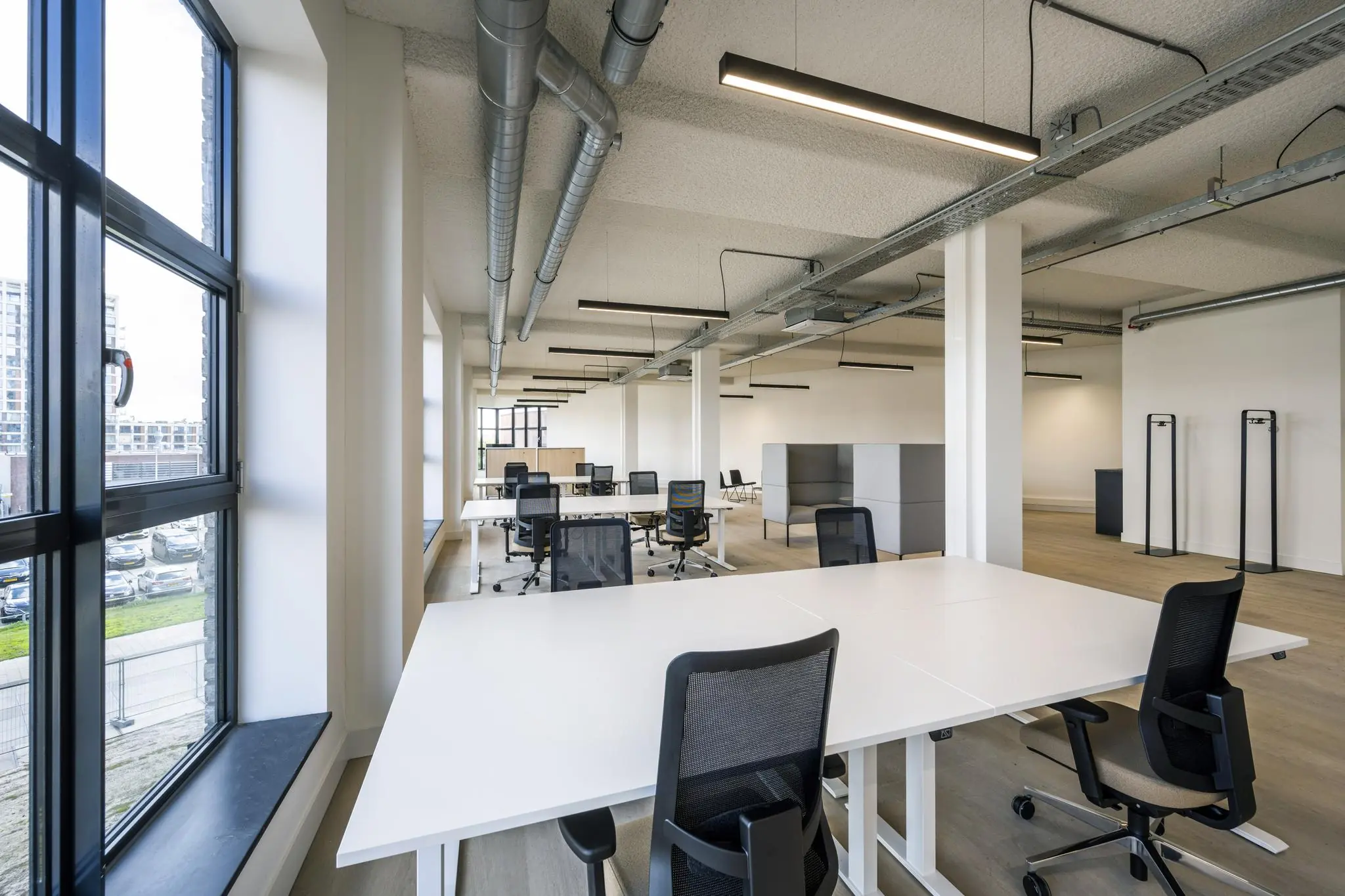 Moderne, open kantoorruimte met witte bureaus, zwarte bureaustoelen en grote ramen aan de linkerkant.