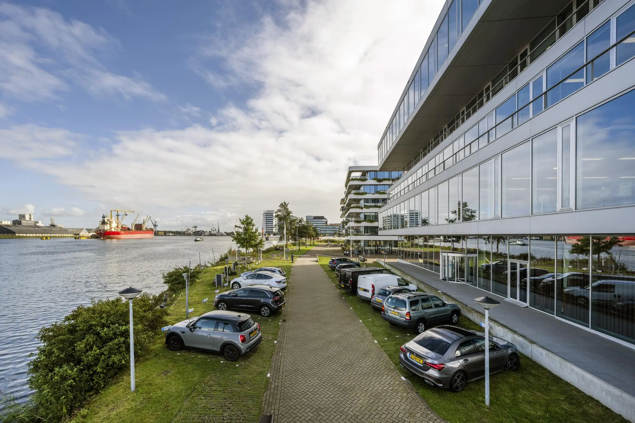 Moderne kantoorgebouwen aan de Moermanskkade met geparkeerde auto's langs het water en een rood vrachtschip op de achtergrond.