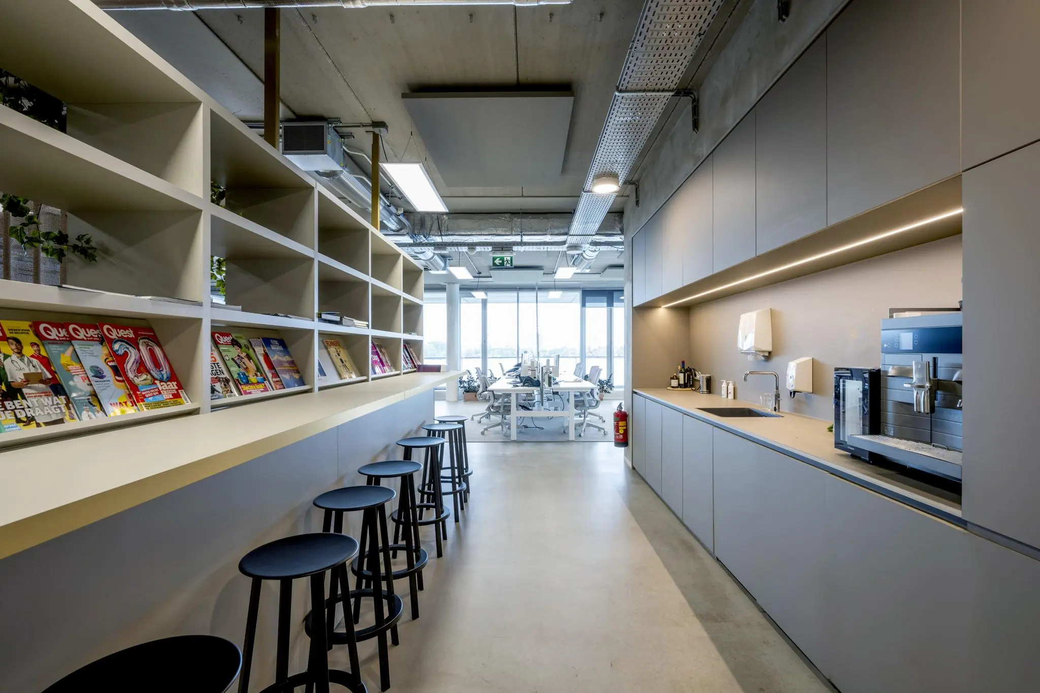 Moderne kantoorpantry aan de Moermanskkade met een koffiehoek, barkrukken, tijdschriftenrek en zicht op een open werkruimte.