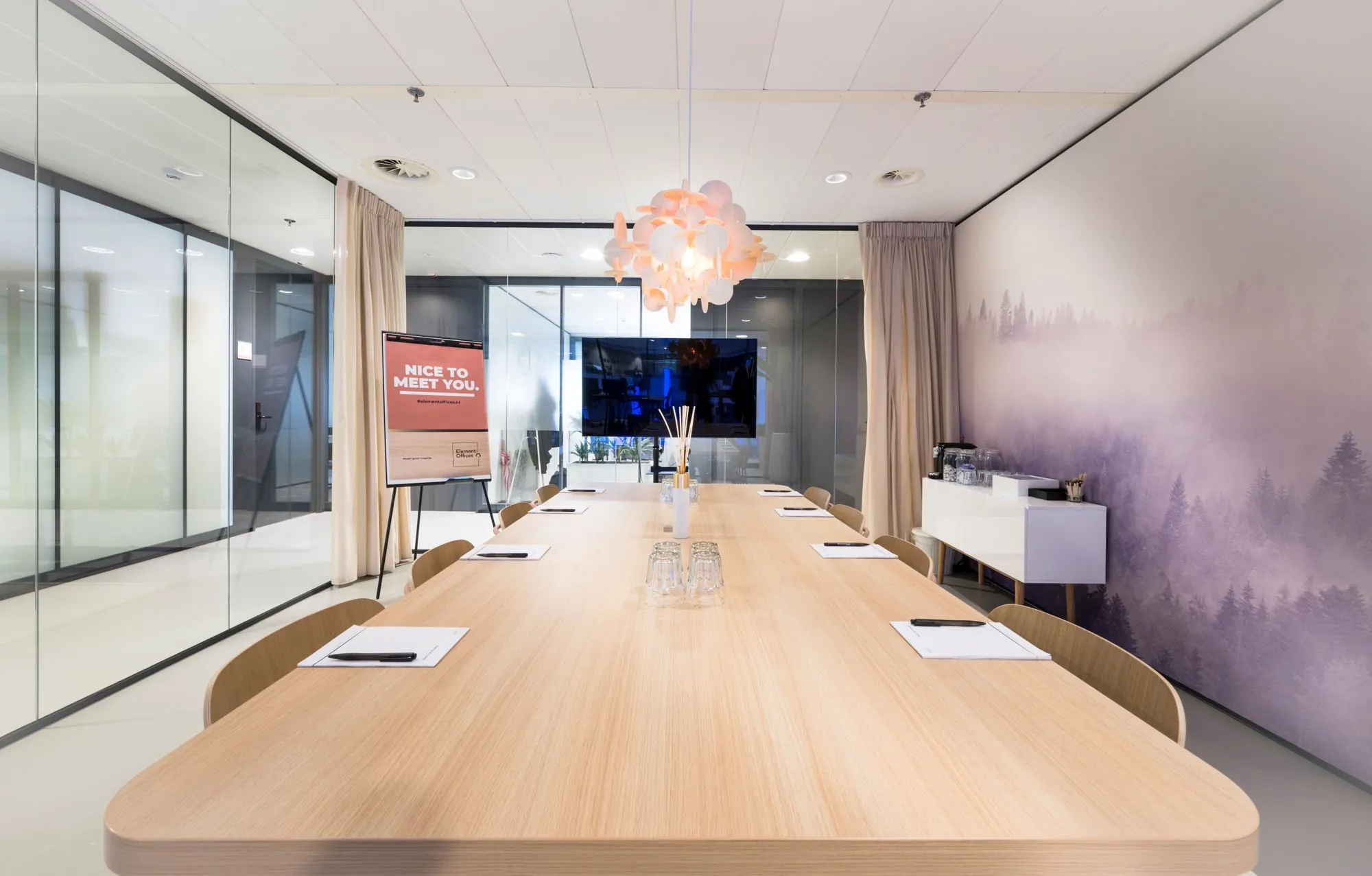Moderne vergaderruimte met een lange houten tafel, notitieblokken, een decoratieve hanglamp en een muurprint van een mistig bos.