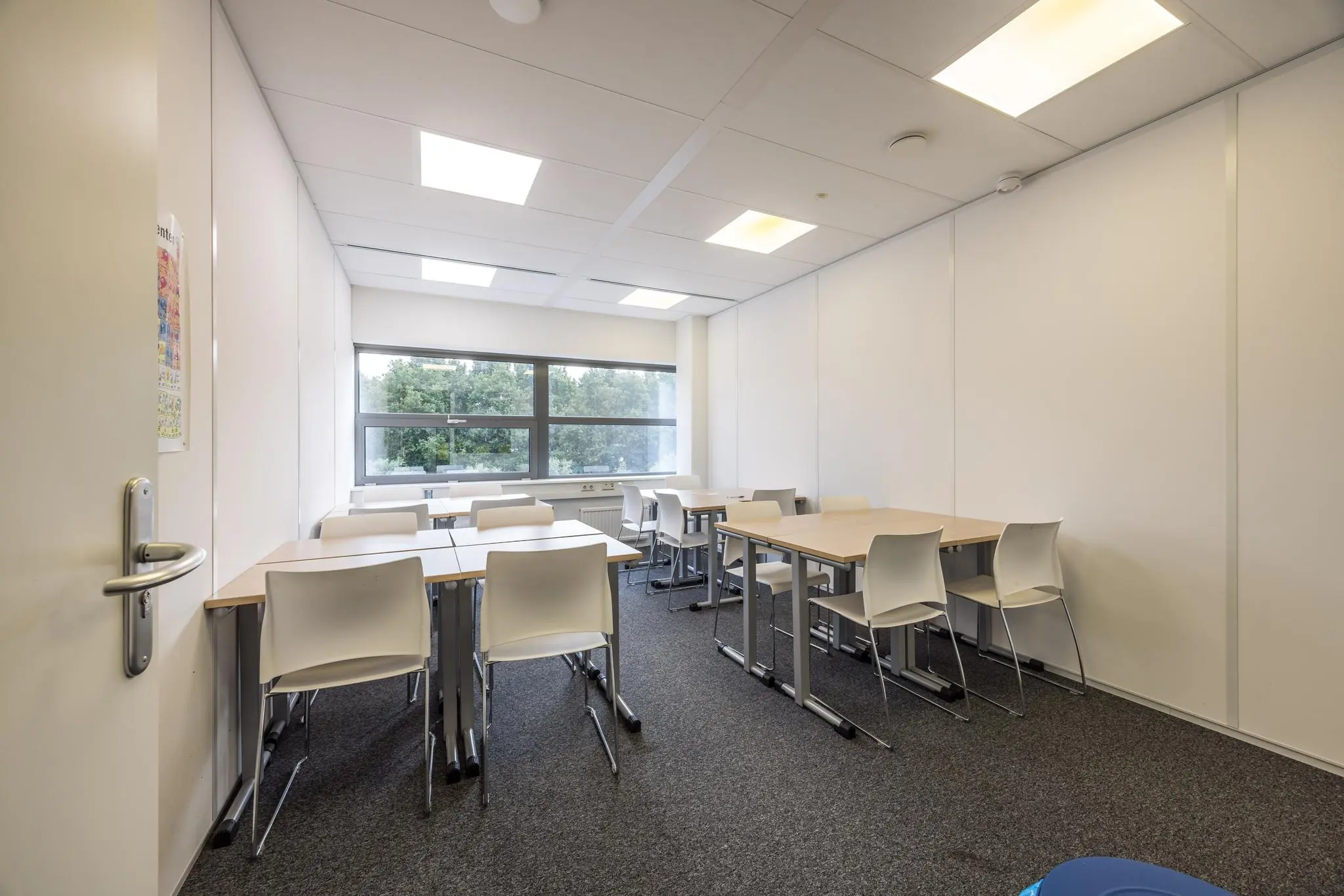 Kleine, moderne vergaderruimte of klaslokaal aan de Daltonlaan met witte muren, bureaus, stoelen en een groot raam met uitzicht op groen.
