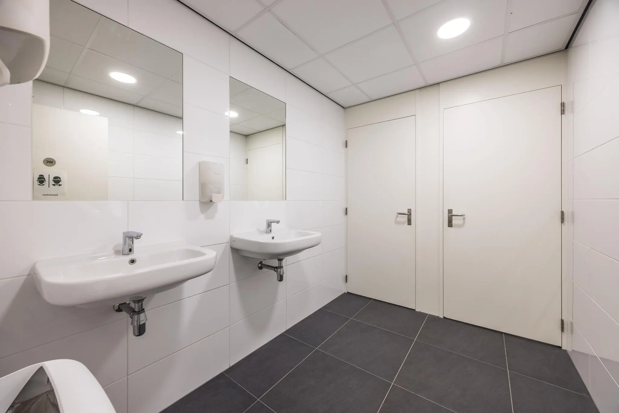 Moderne, schoon ogende openbare toiletruimte met twee wastafels, spiegels en deuren naar toiletcabines in een gebouw aan de Daltonlaan.