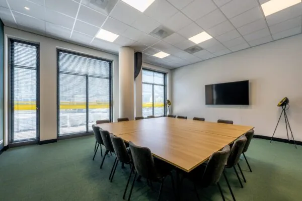 Vergaderruimte aan het Elizabeth Cady Stantonplein met grote vierkante tafel, zwarte stoelen, flatscreen aan de muur en hoge ramen met jaloezieën.