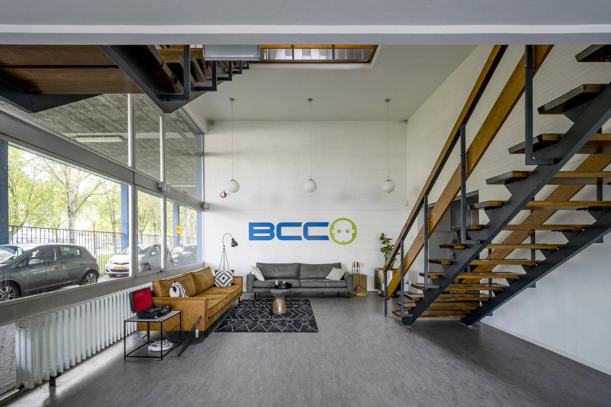 Moderne wachtruimte met BCC-logo, twee banken, een draaitafel en een houten trap in een lichtovergoten interieur.
