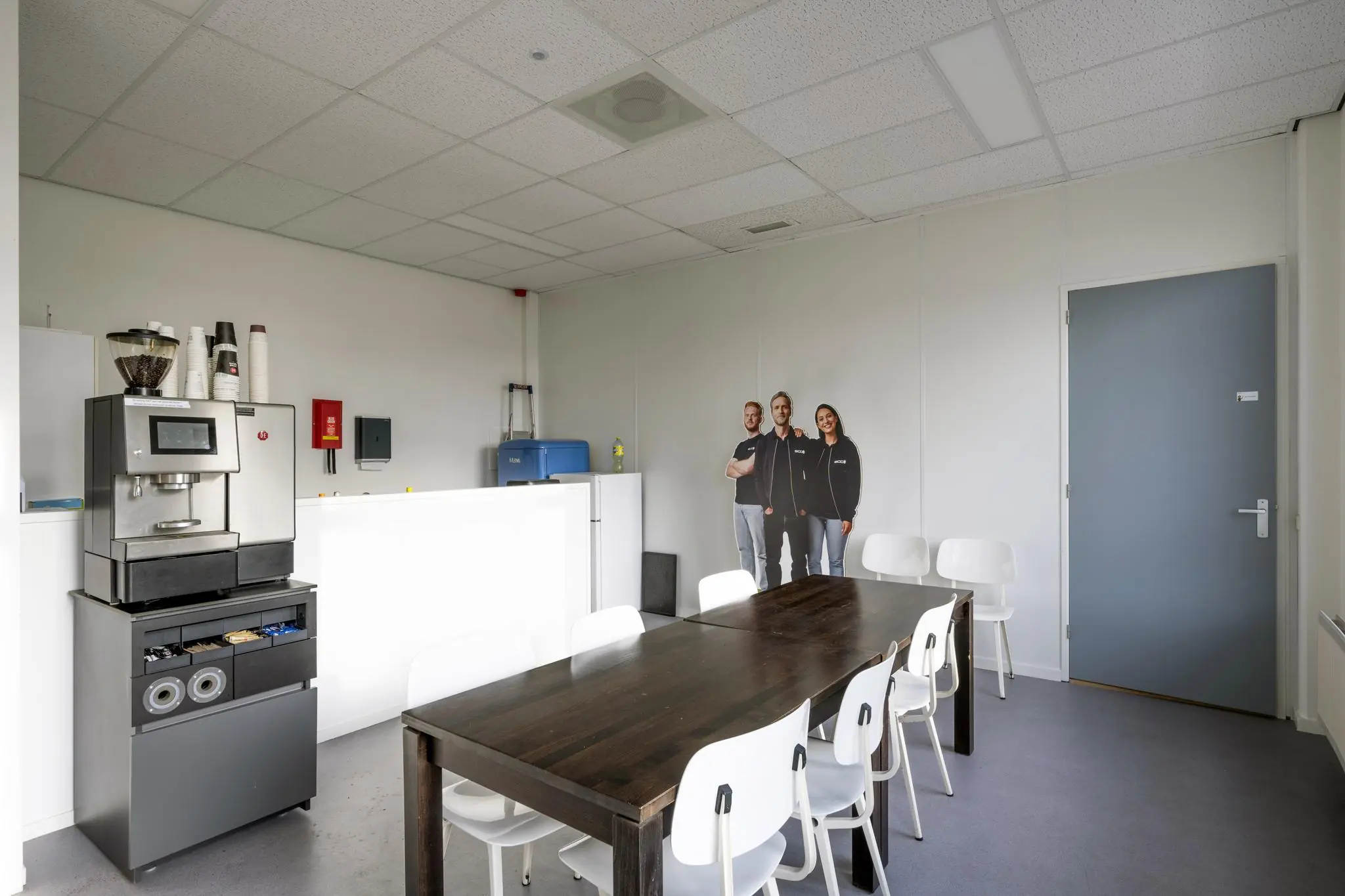 Kantineruimte aan de Van der Madeweg met een koffiemachine, houten vergadertafel en een kartonnen display van drie personen.