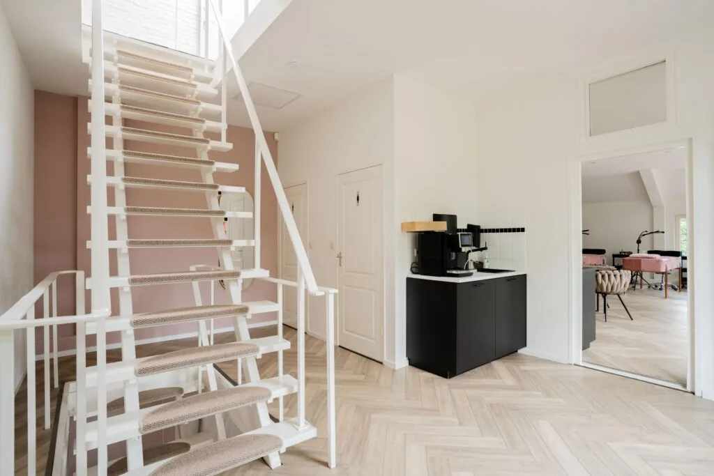 Moderne hal met witte open trap, koffiecorner en zicht op een lichte ruimte met behandelstoelen aan de Sloterweg.
